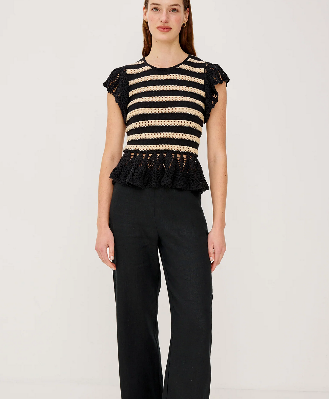 A woman stands against a plain background wearing the Rails Coen Knit Top in black stripe with cap sleeves, paired with black wide-leg pants.