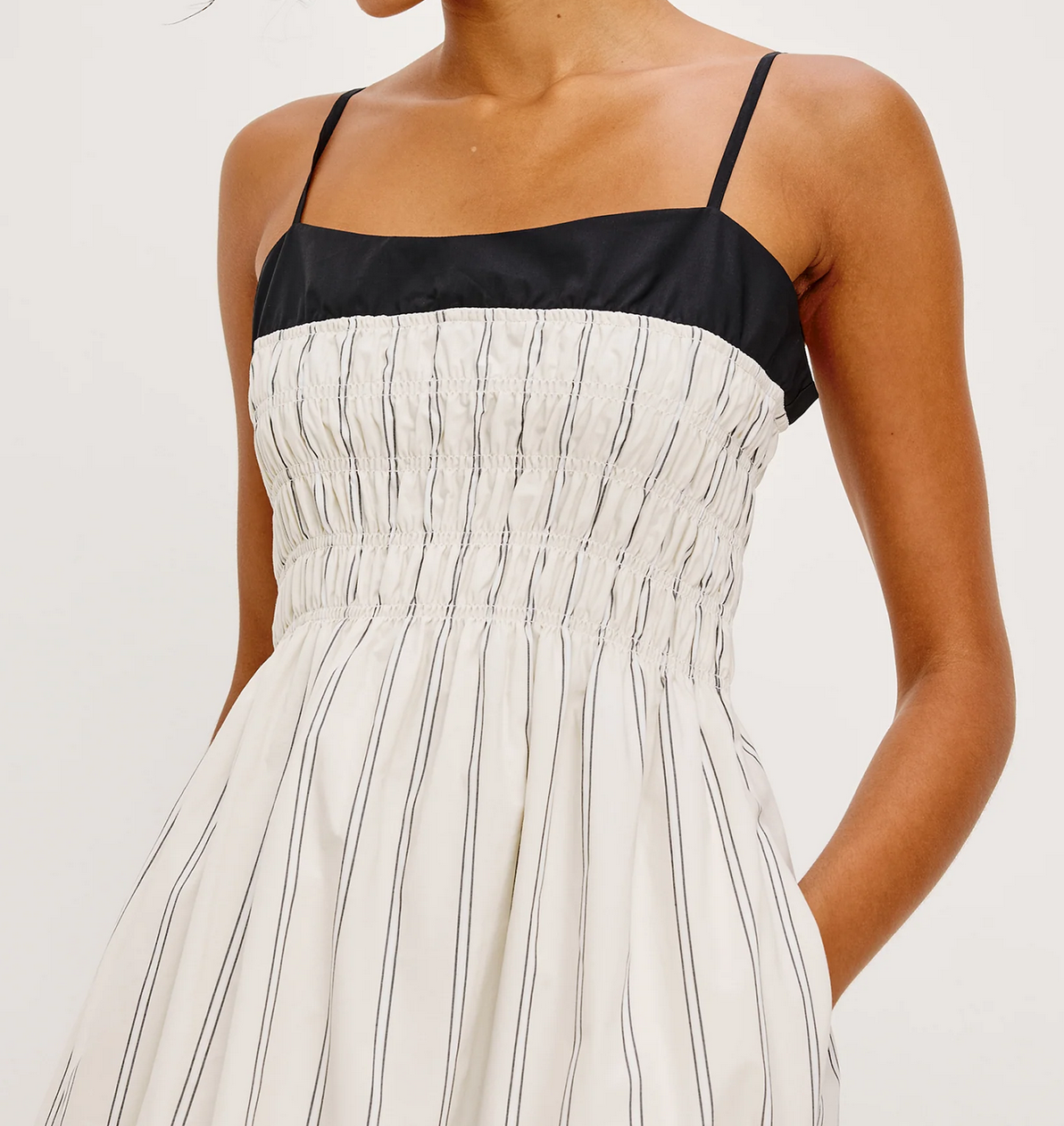 A woman wears the Rails Primrose Dress in Ecru Stripe, featuring thin straps, a black bodice, and a white skirt with vertical black stripes and gathered waist detailing.