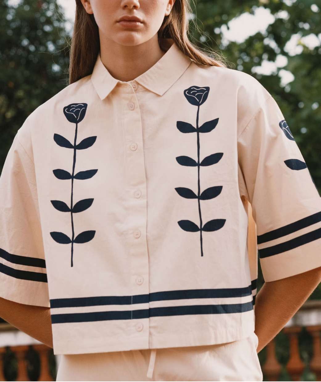 Woman wearing a beige shirt with black floral patterns and a matching hat outdoors.