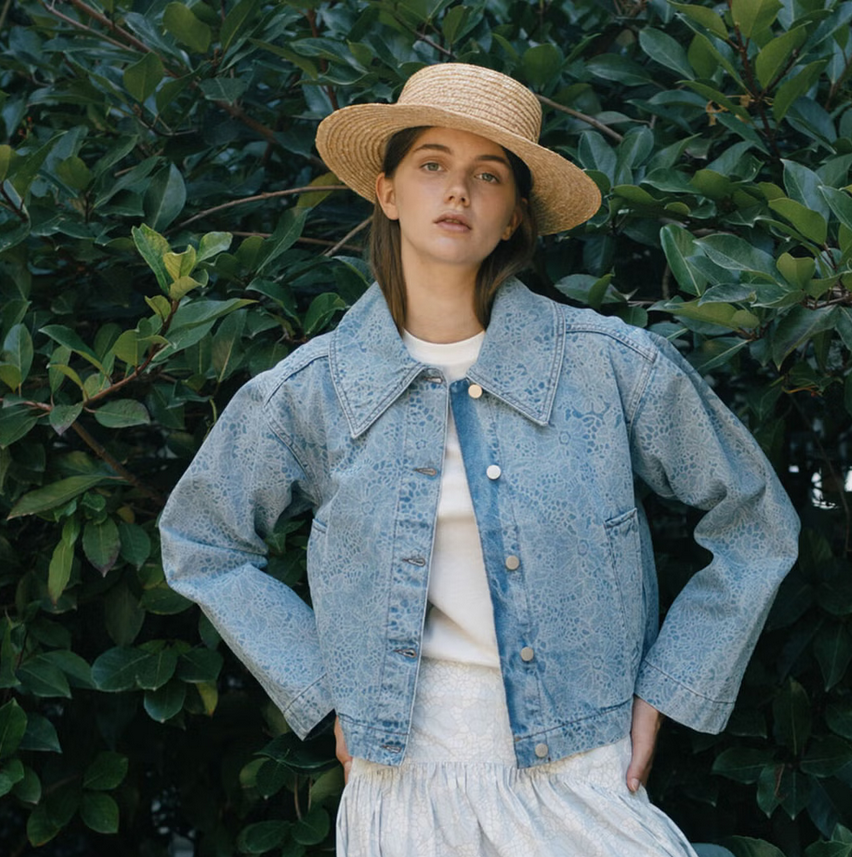 A woman stands before leafy bushes wearing the Tiny Big Sister Laser Lace Denim Jacket in light blue, a white skirt with laser-cut lace, and a straw hat.