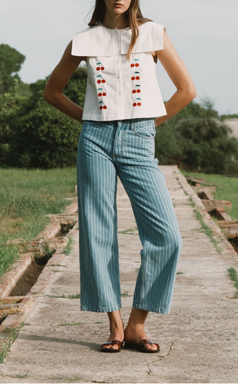 Outdoors on a concrete path, a person wears a white sleeveless top with red and blue details, brown sandals, and Tiny Big Sister's Berta Wide Leg Jeans - Blue Stripe.