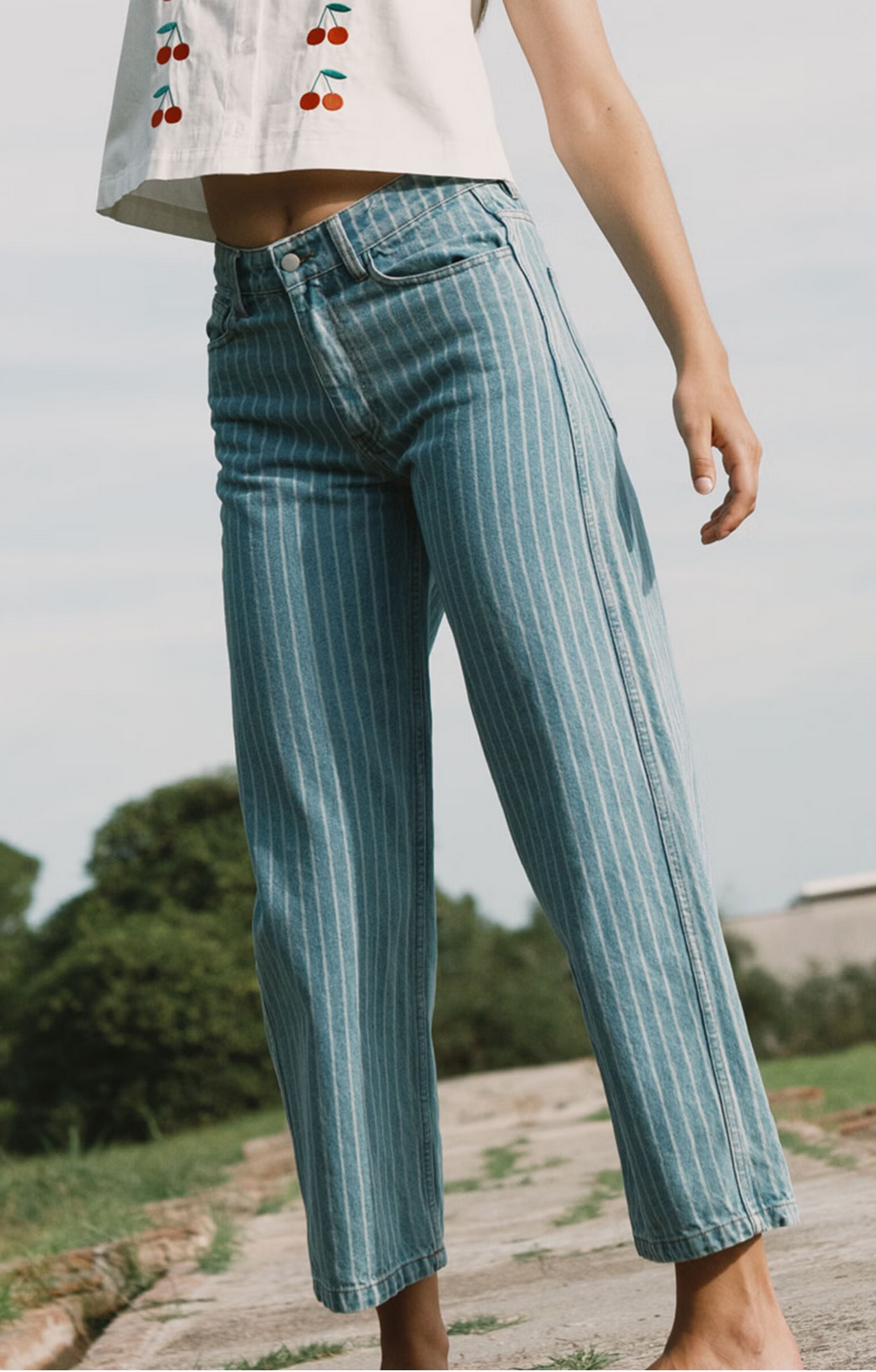 A person stands outdoors in the Tiny Big Sister Berta Wide Leg Jeans - Blue Stripe and a white cherry-embroidered crop top, surrounded by greenery and sky.