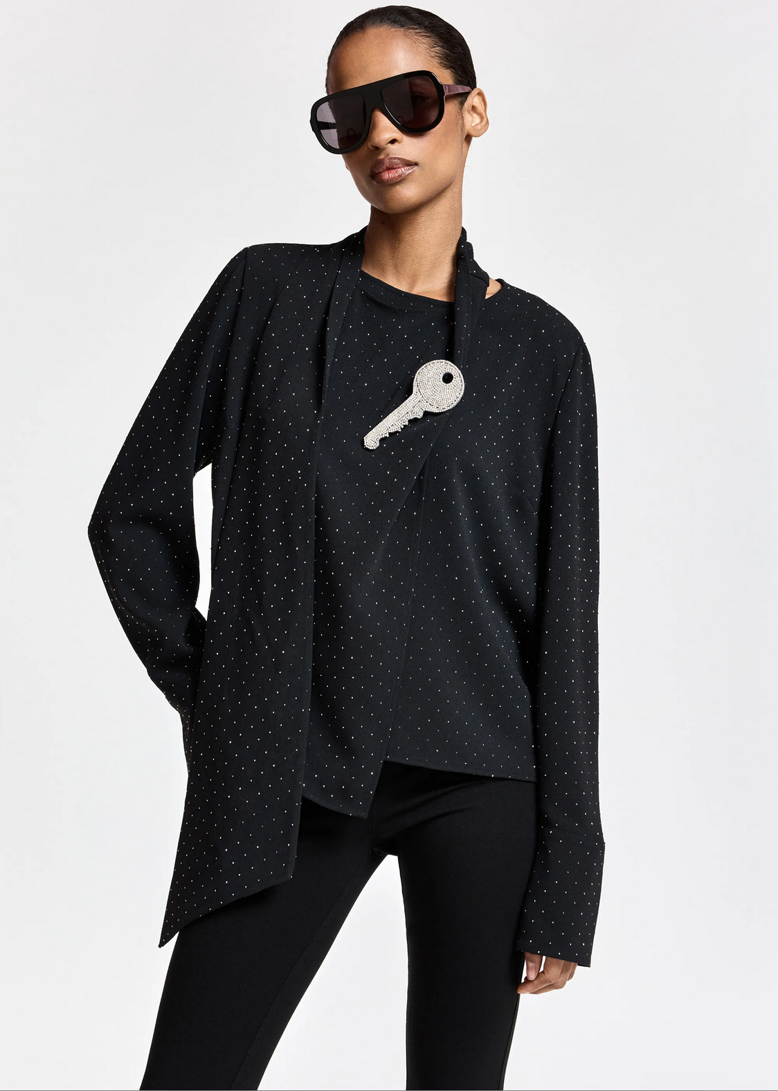 A person wears the Essentiel Antwerp Kalanchoe Top with Scarf—featuring black dotted fabric and asymmetric panels—paired with black pants, large dark sunglasses, and a necklace with an oversized silver key pendant, standing against a plain light background.
