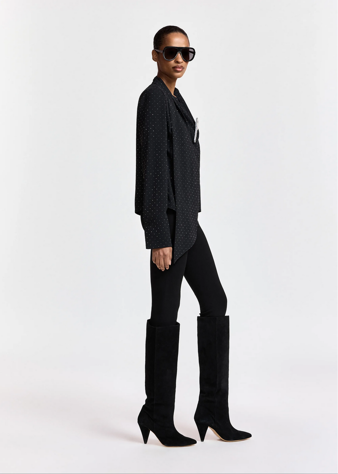A woman stands in profile against a white background, wearing sunglasses, the Essentiel Antwerp Kalanchoe Top with Scarf featuring a black polka-dot pattern and rhinestone embellishments, paired with black leggings and black knee-high heeled boots.