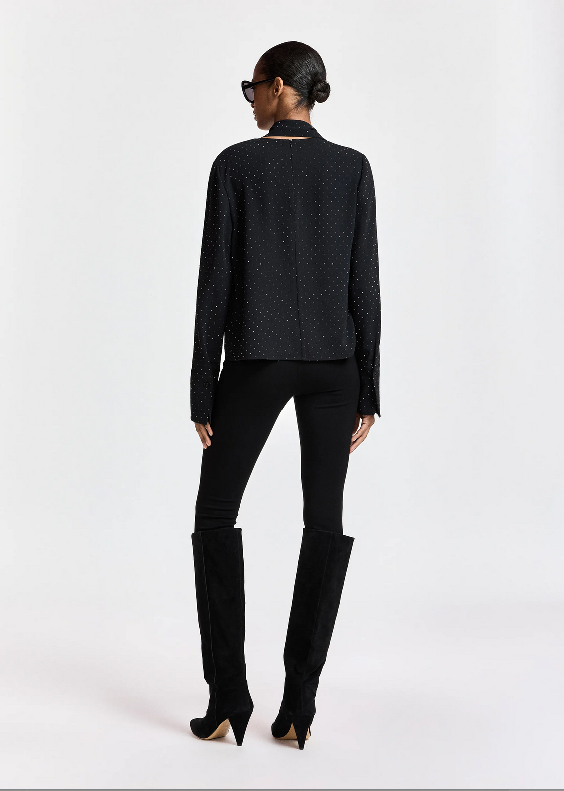 A person is seen from behind, wearing the Essentiel Antwerp Kalanchoe Top with Scarf—a black long-sleeve top featuring an asymmetric panel—paired with black pants and black knee-high boots, standing against a plain white background.