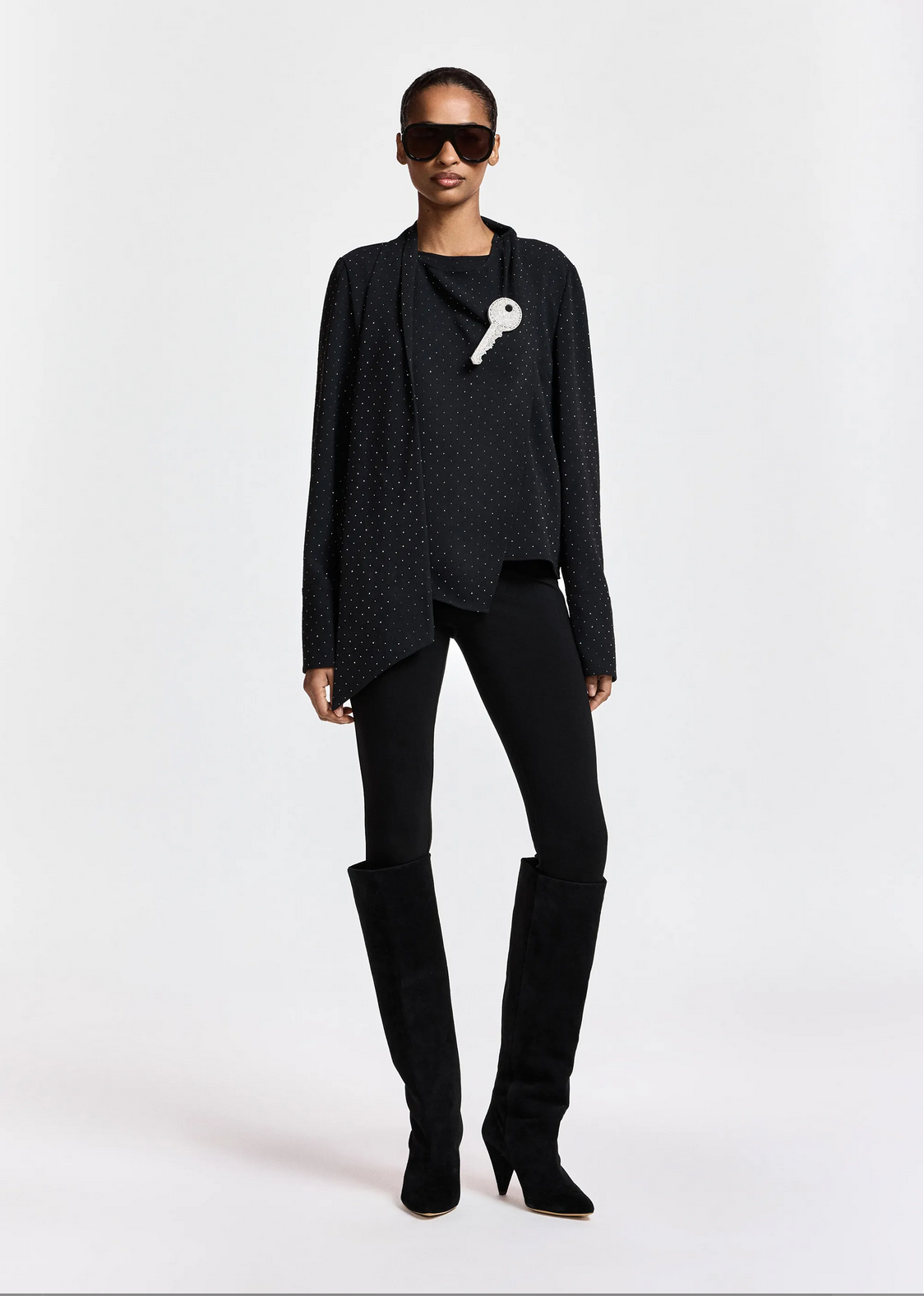 A person is wearing the Essentiel Antwerp Kalanchoe Top with Scarf, styled in an all-black ensemble beneath a polka-dotted jacket, paired with black leggings, knee-high boots, oversized sunglasses, and a large decorative key pinned to the jacket, standing against a plain background.