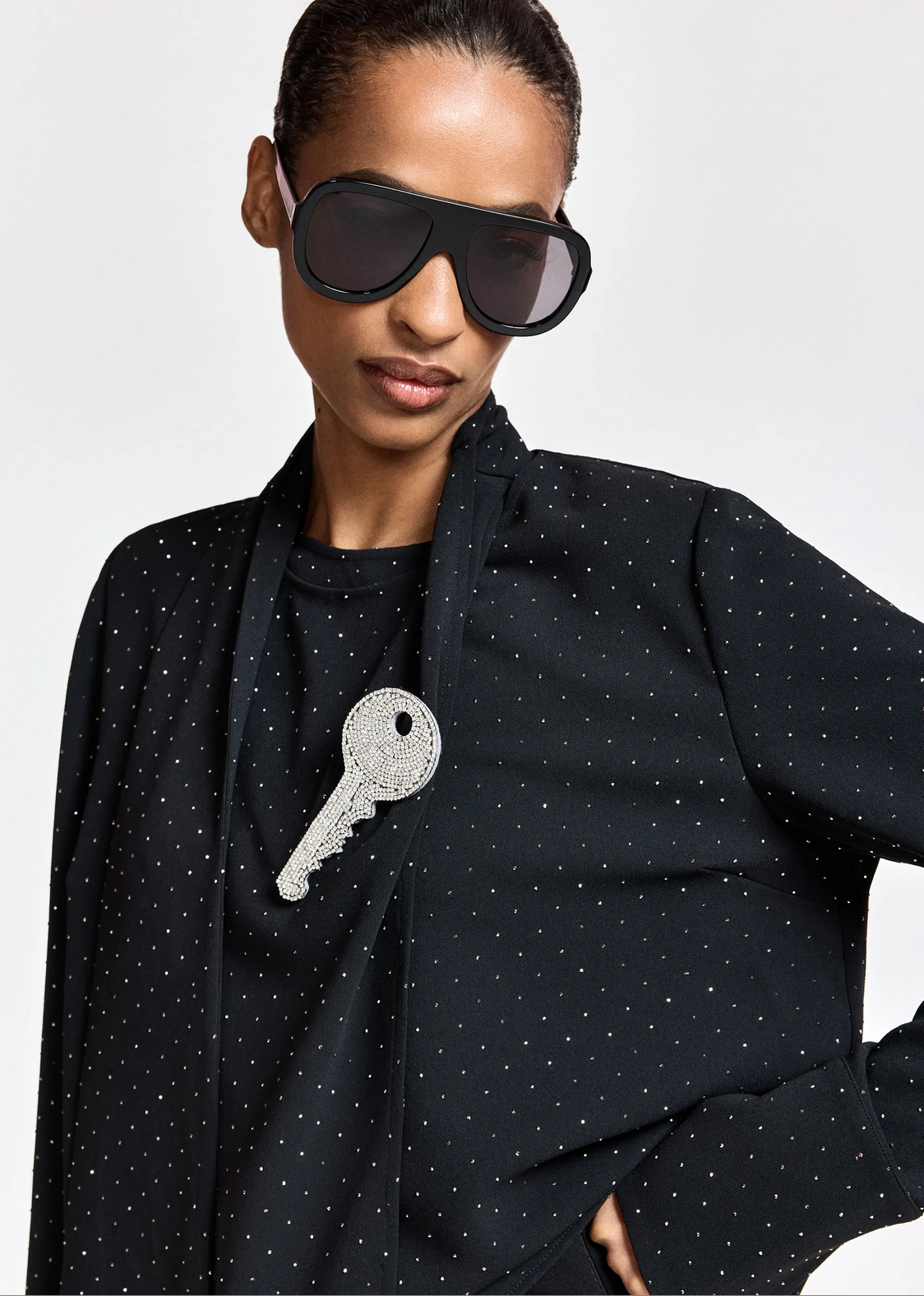A person wearing large black sunglasses and the Essentiel Antwerp Kalanchoe Top with Scarf—a black polka dot style with a draped neckline—accented by a sparkling key-shaped pendant necklace and subtle rhinestone embellishments.