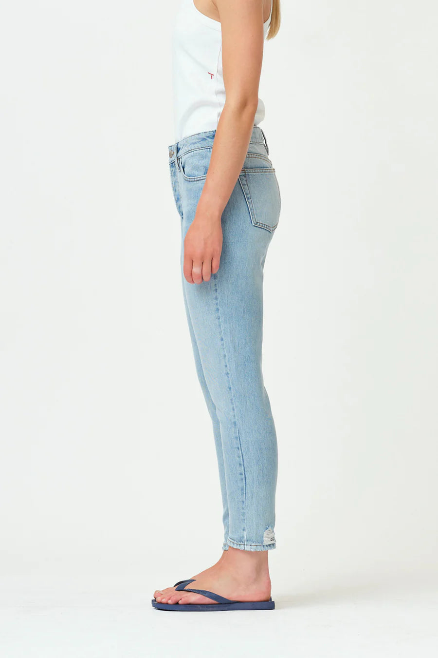 A woman in a white tank top and Hepburn Slim Jeans - Versailles Dist. by Tomorrow Denim, standing in front of a white wall.