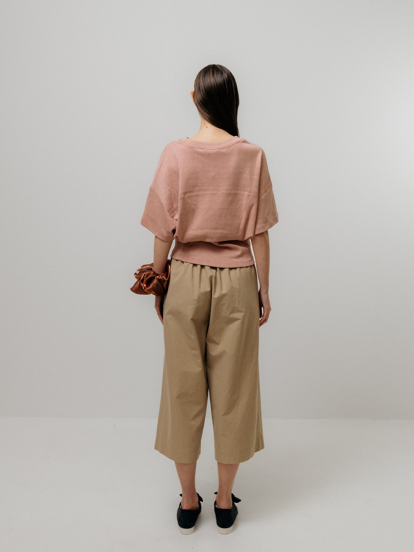 A person with their back to the camera wears Emin and Paul's Tapered T-Shirt in soft pink, paired with beige wide-leg pants and black shoes while holding a brown scrunchie against a plain background.