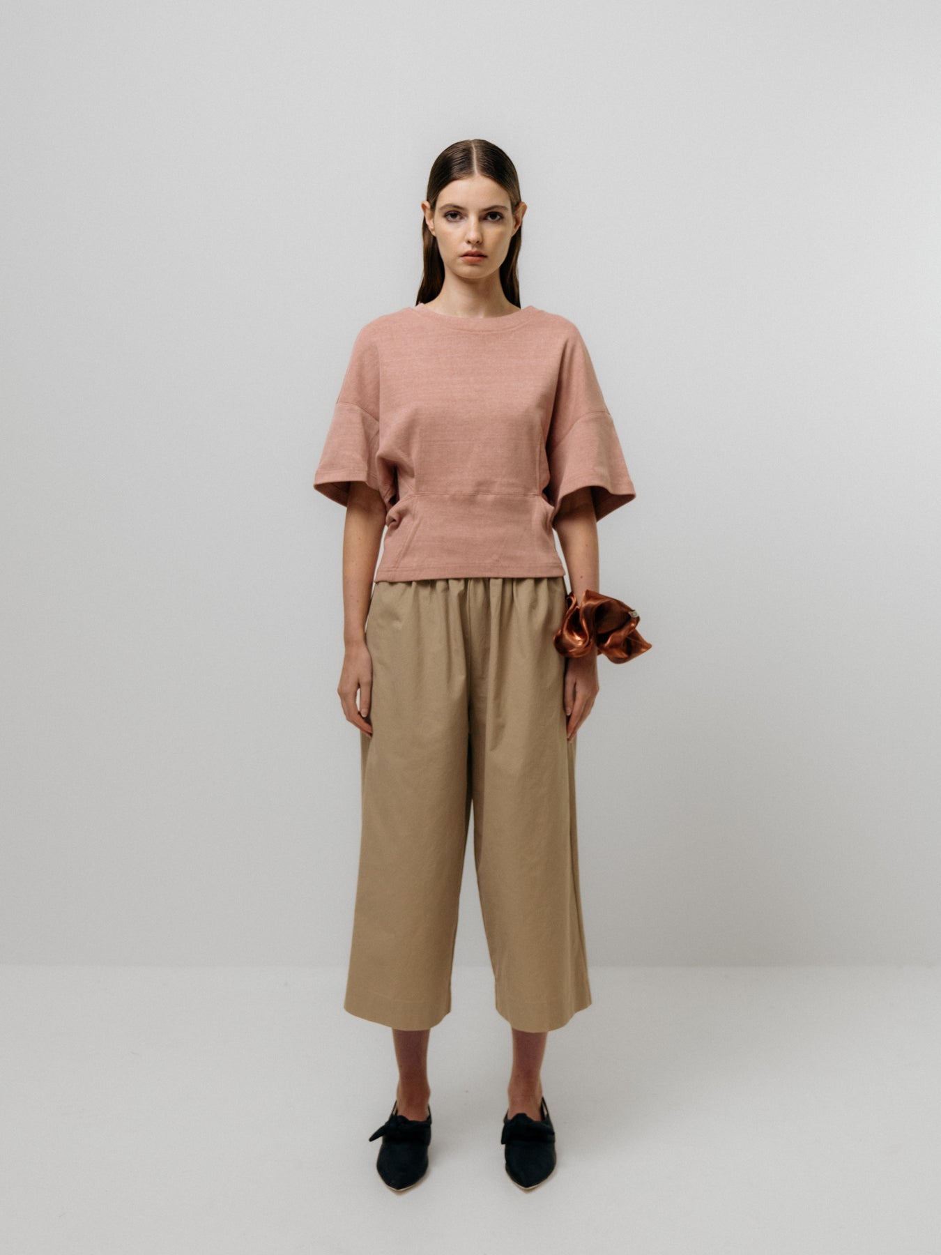 A woman wears a Tapered T-Shirt in soft pink by Emin and Paul, tan wide-legged pants with a structured waist, and black shoes against a neutral background, holding a rust-colored accessory.