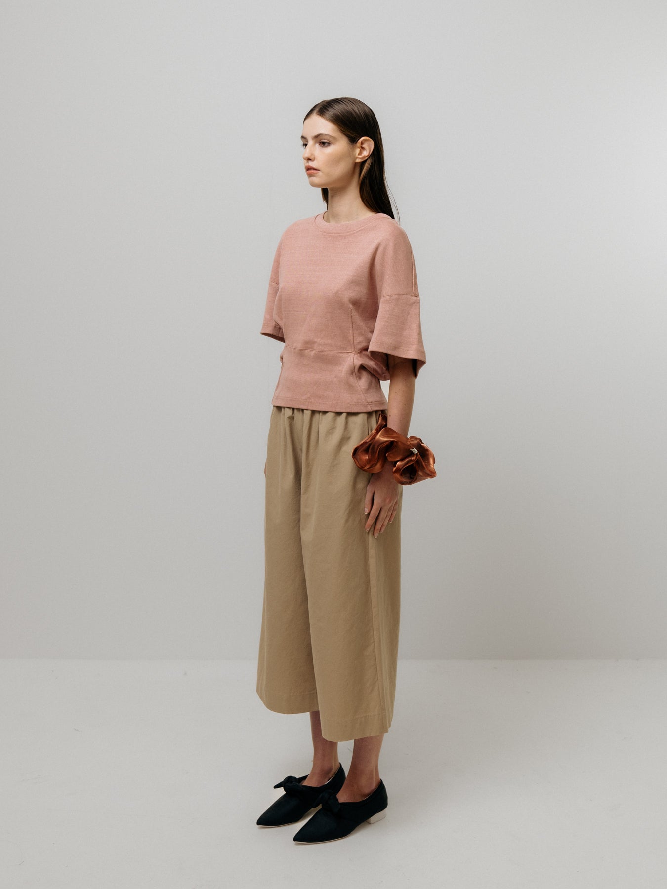 A woman with long hair stands against a plain background in Emin and Paul's Tapered T-Shirt - Pink, made from 100% cotton, beige wide-leg pants featuring a structured waist, and black shoes. She wears a large scrunchie on her wrist.