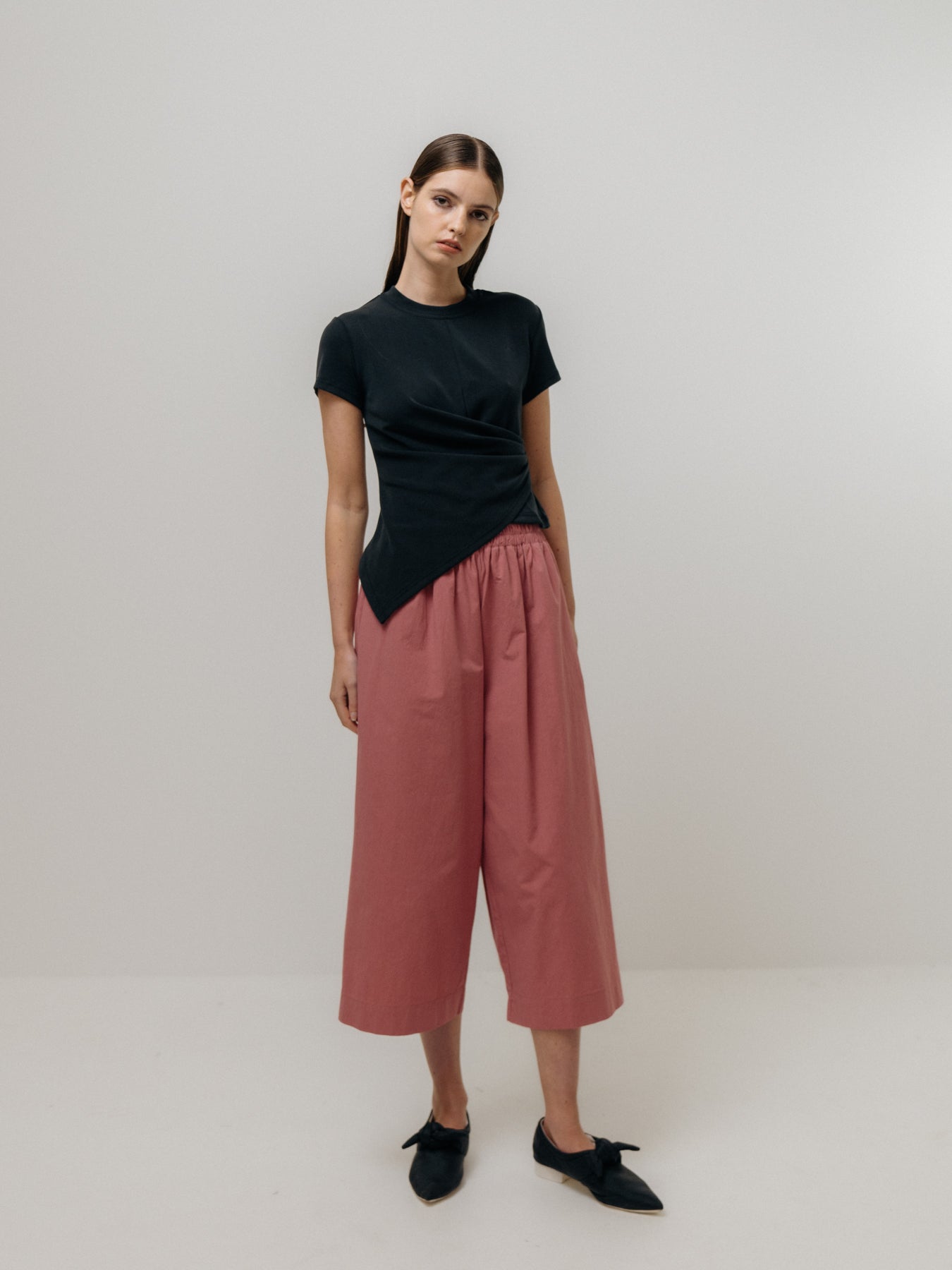 A person stands against a plain background wearing a black asymmetrical top, Cotton Culottes in Pink by Emin and Paul, and black shoes.