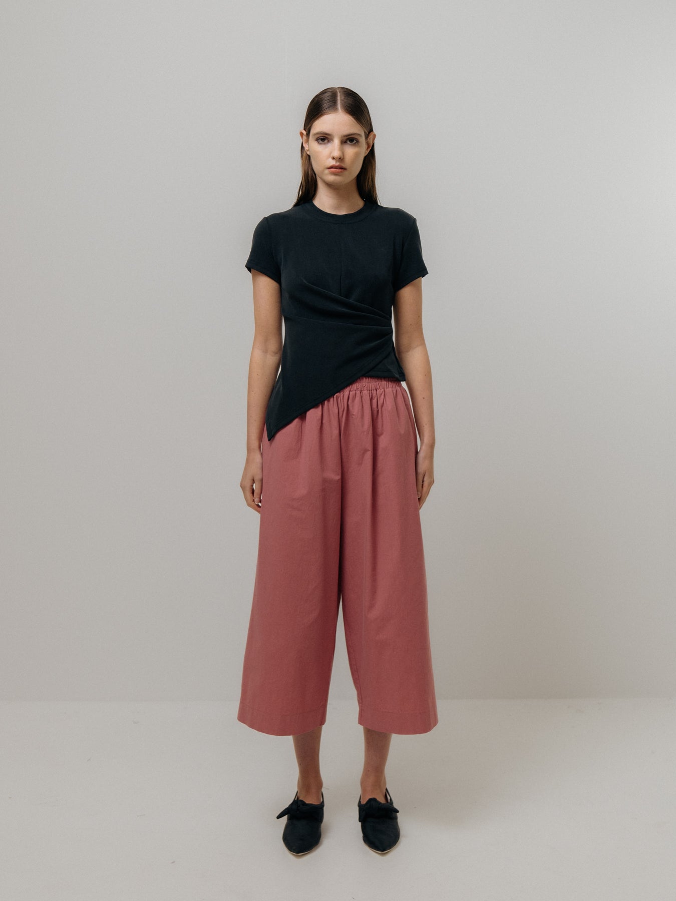 A person poses against a plain backdrop in a black asymmetrical top, Cotton Culottes - Pink by Emin and Paul, and sleek black shoes.