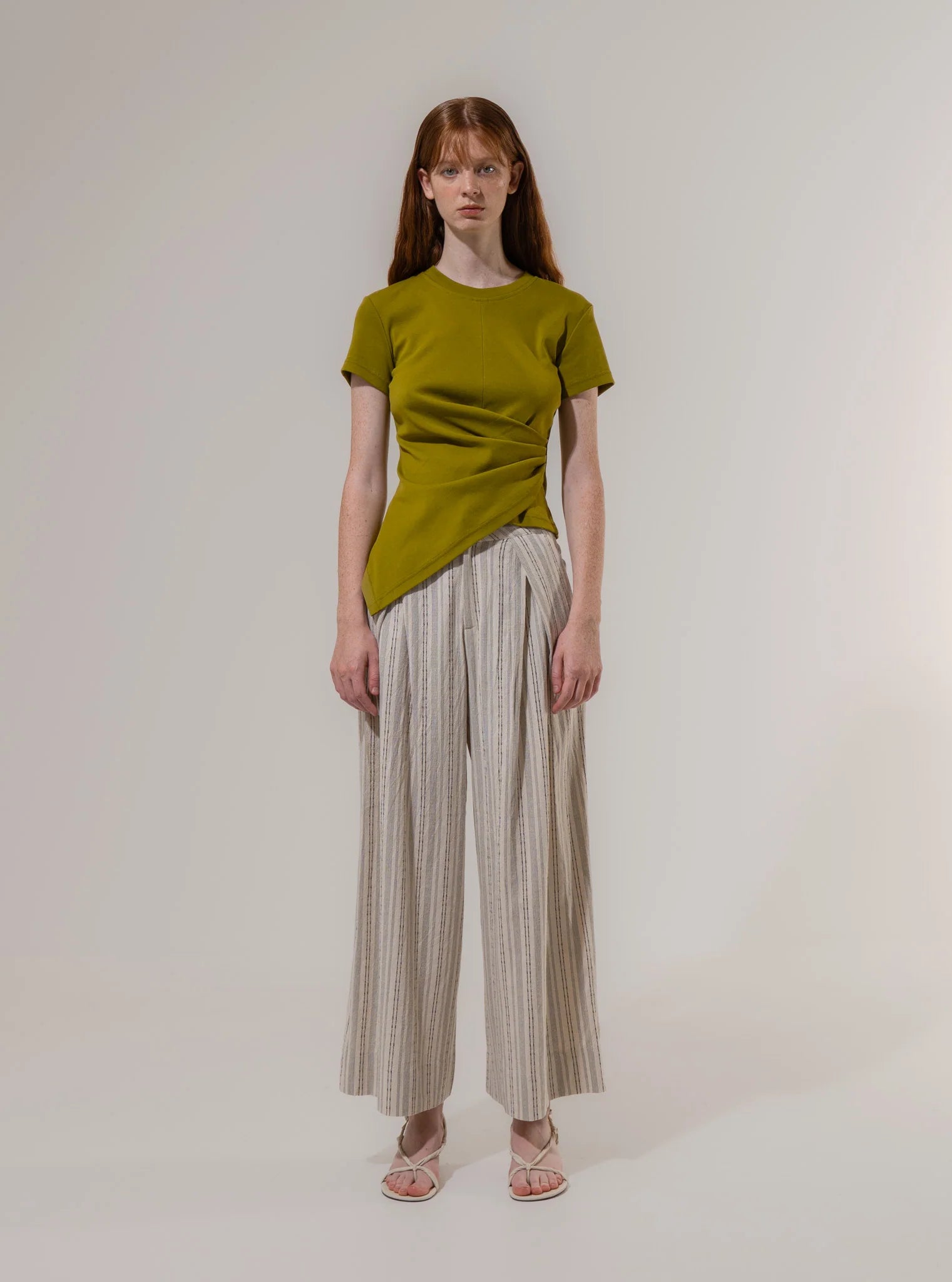 A woman stands against a plain background wearing a green short-sleeve top, Emin and Paul Structured Pleat Trousers in Cream Stripe with wide legs, and sandals.