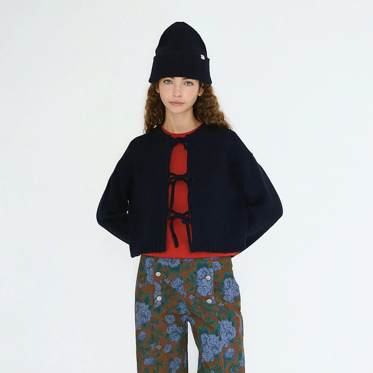 A woman wears the Tiny Big Sister Tie Detail Cardigan in navy, paired with a red top, floral brown pants, and a navy knit beanie while standing against a plain white background.