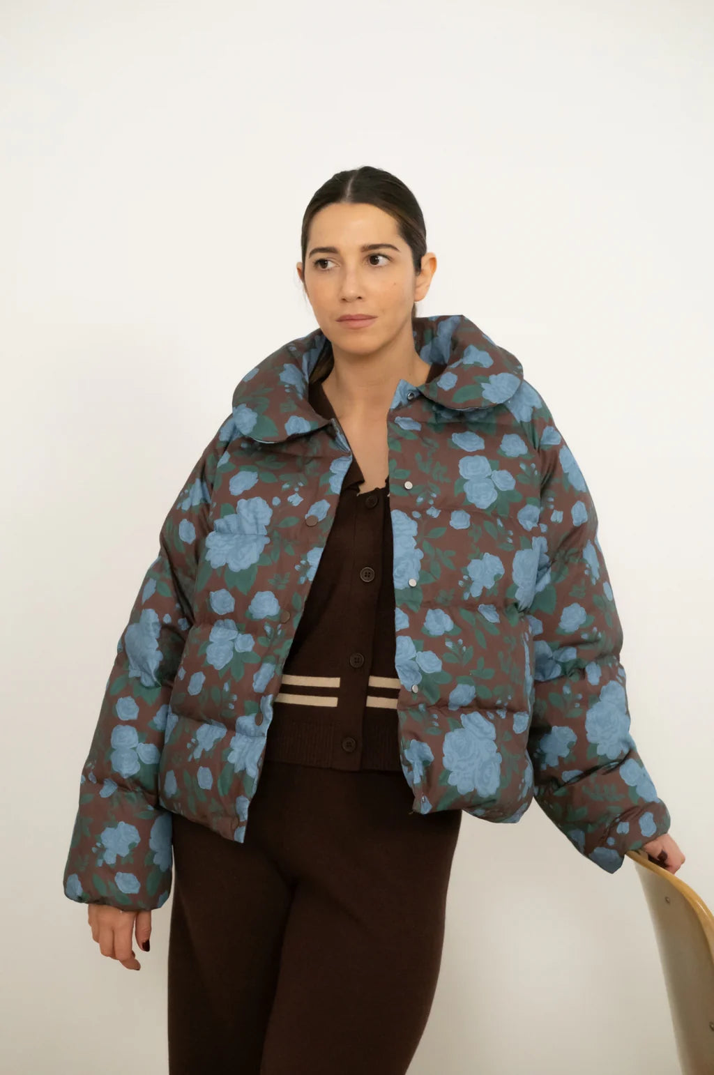 A woman stands against a plain background wearing the Tiny Big Sister Roses Short Puffer Jacket in Brownn, styled with a brown cardigan and pants.