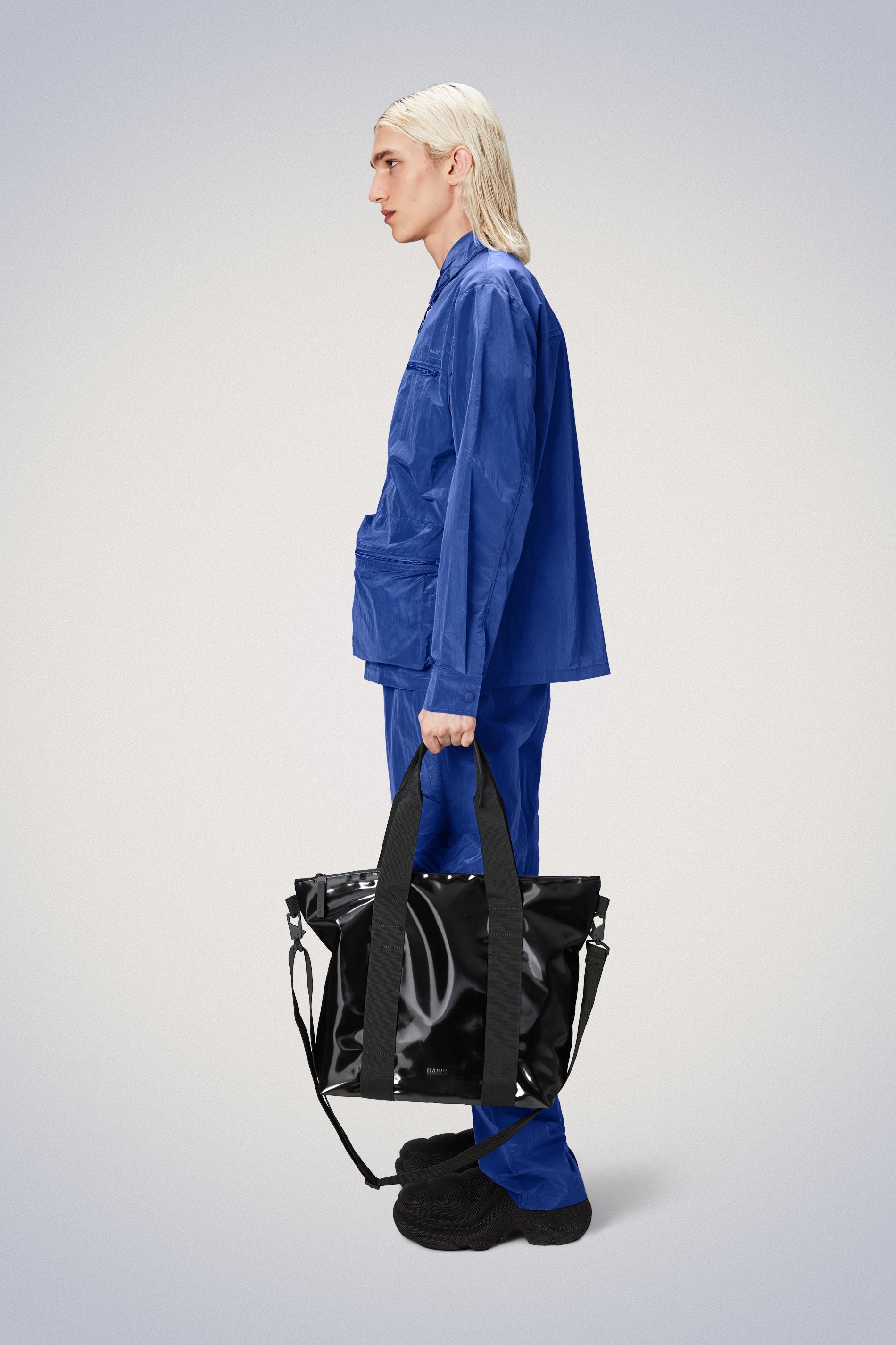 A woman in blue pajamas holding a Rains Tote Bag Mini - Night made from durable materials, featuring reinforced stitching for added durability. The mini size provides plenty of storage space while being easy to carry.