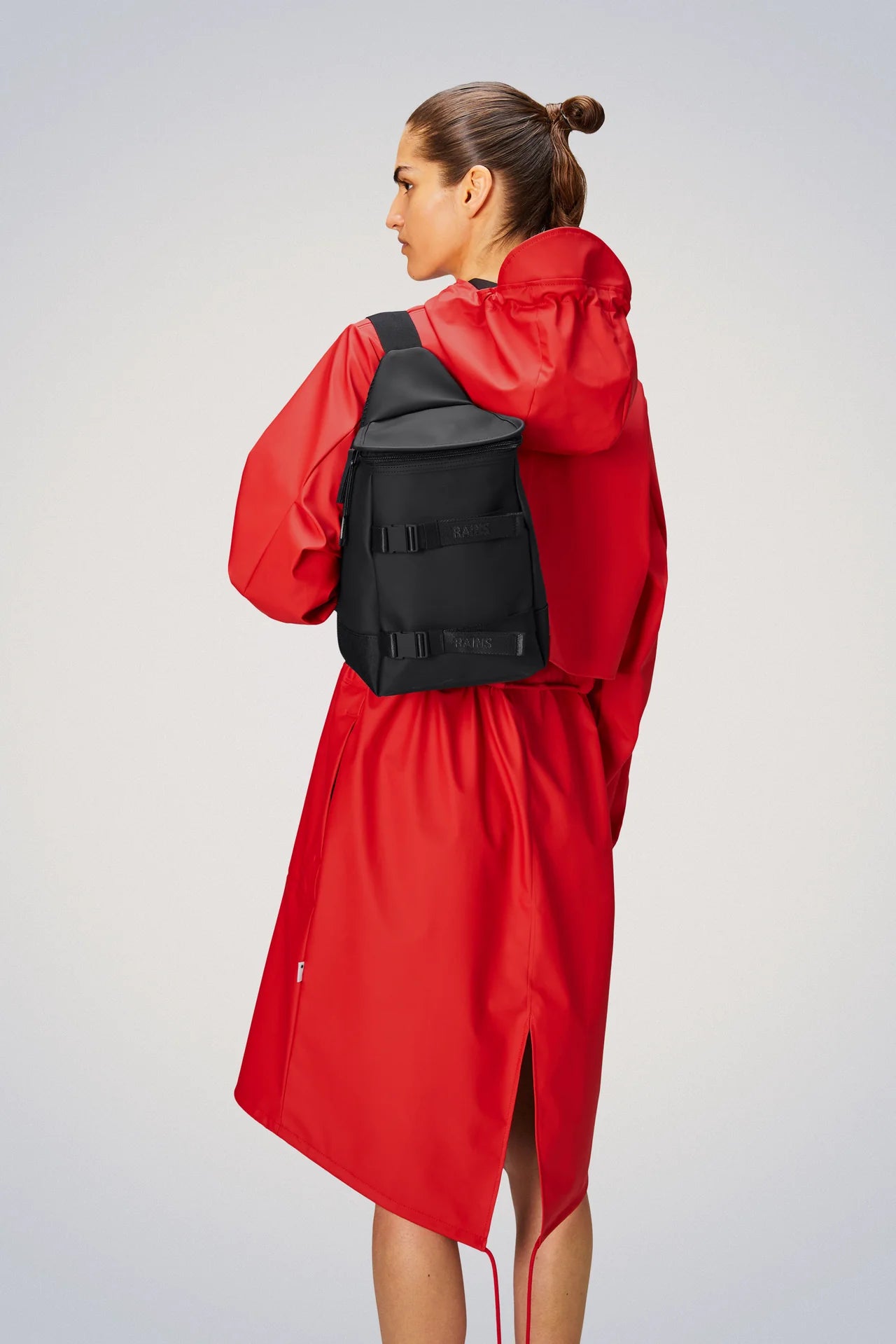A woman in a waterproof red raincoat with a Rains Trail Sling Bag - Black slung over her shoulder.