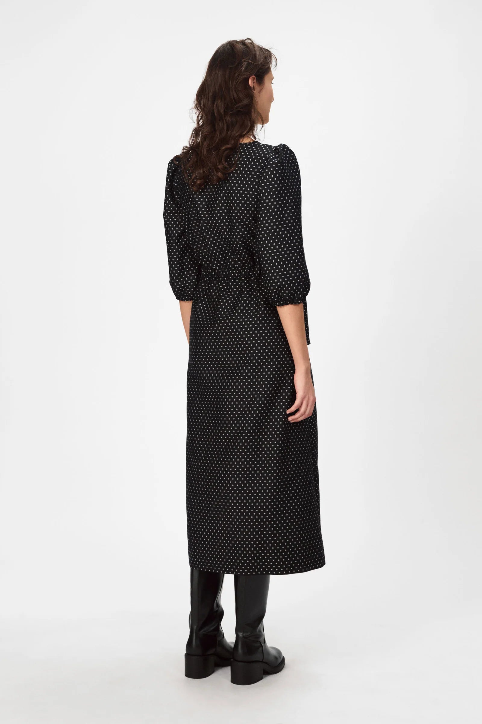 A person with long curly hair wears the Nué Notes Vilnius Dress in Black Polkadot, paired with black boots, standing facing away against a plain white background.