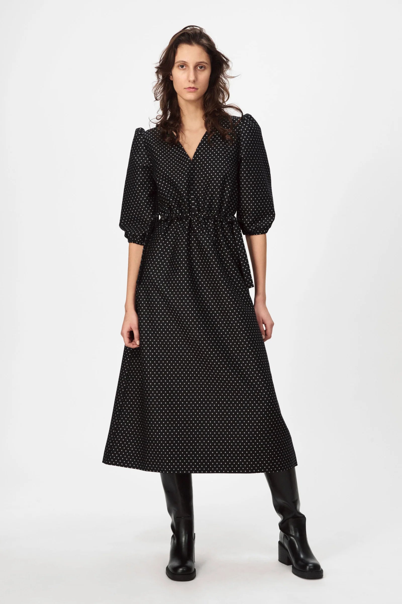 A person wearing the Nué Notes Vilnius Dress in Black Polkadot and black knee-high boots stands against a plain white background.