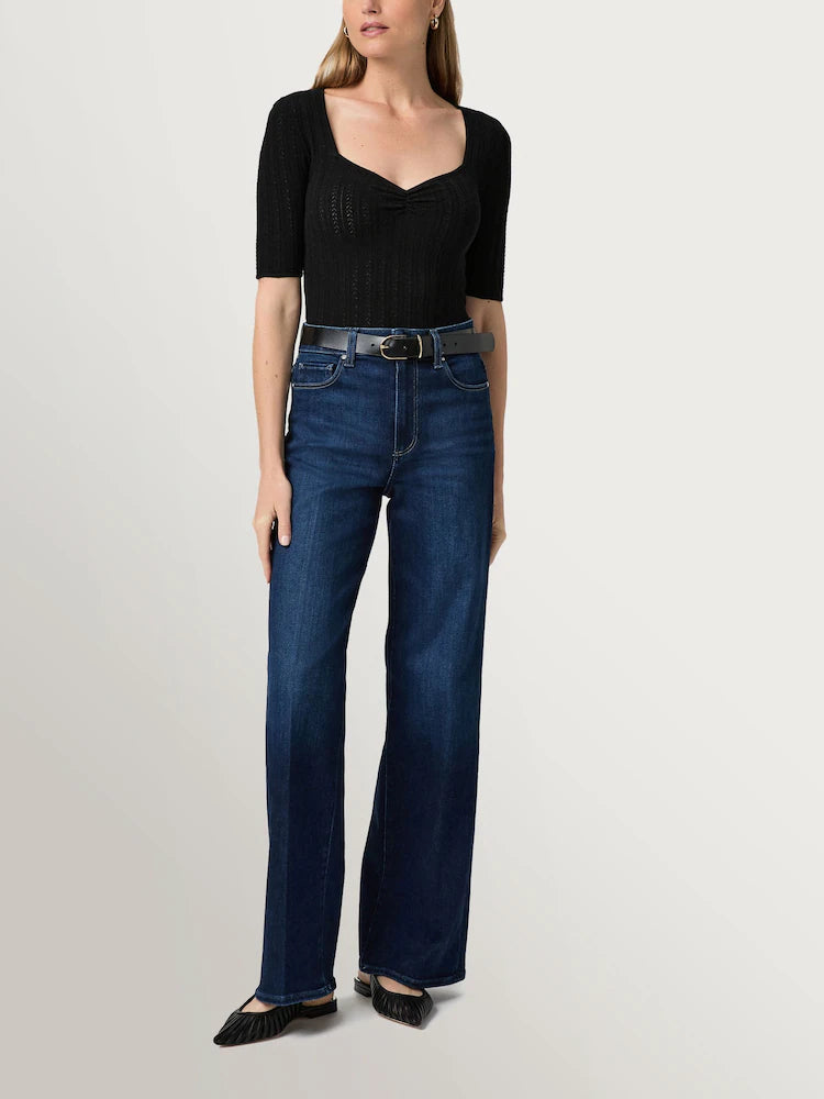 Woman wearing a fitted black short-sleeve top, Paige Anessa Wide Leg 31"- Duke of York jeans with a black belt, and black pointed shoes, standing against a plain light background.