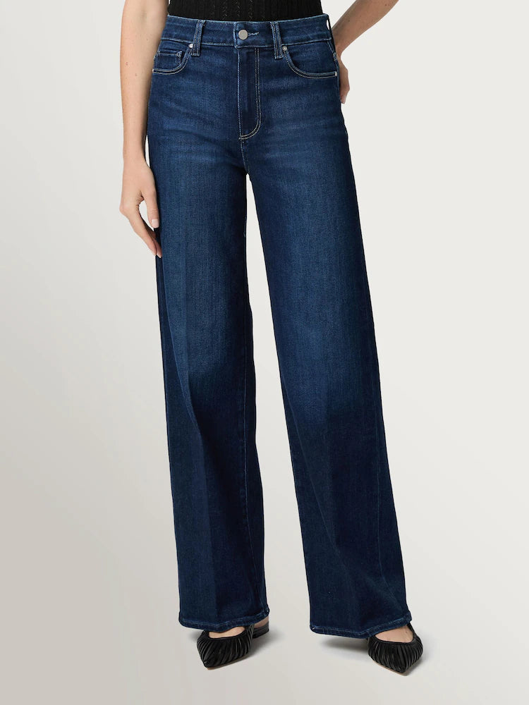 A person wears Paige Anessa Wide Leg 31"- Duke of York jeans in dark blue vintage denim paired with black pointed shoes, standing against a light background.