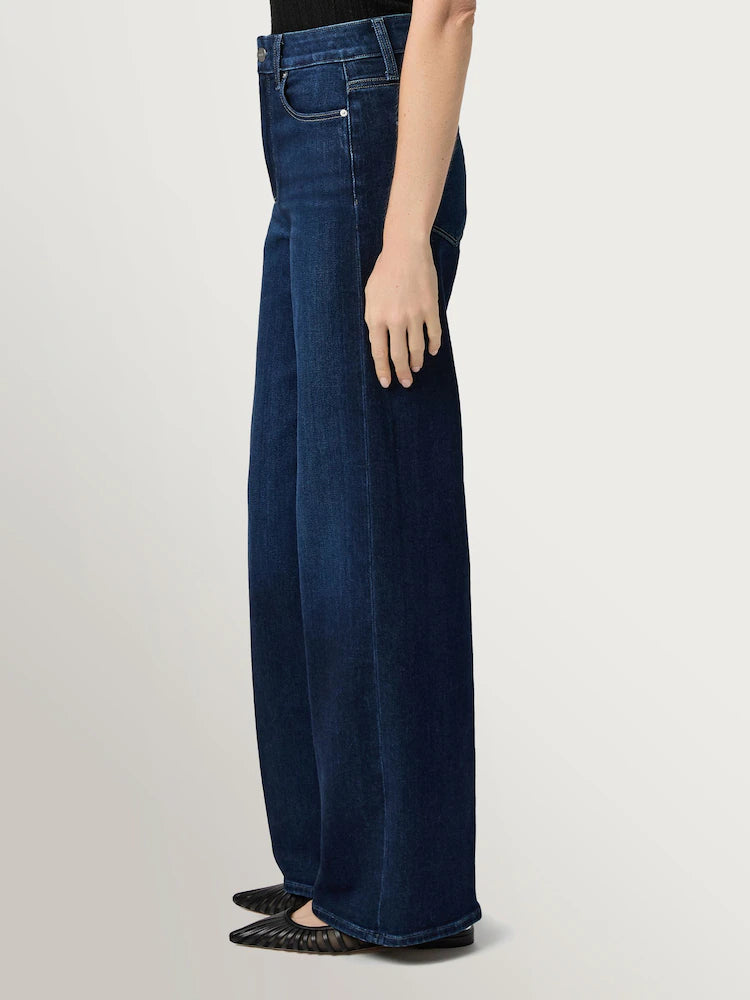 A person stands in profile against a plain background wearing Paige's Anessa Wide Leg 31"- Duke of York jeans in dark blue vintage denim and black pointed-toe shoes.
