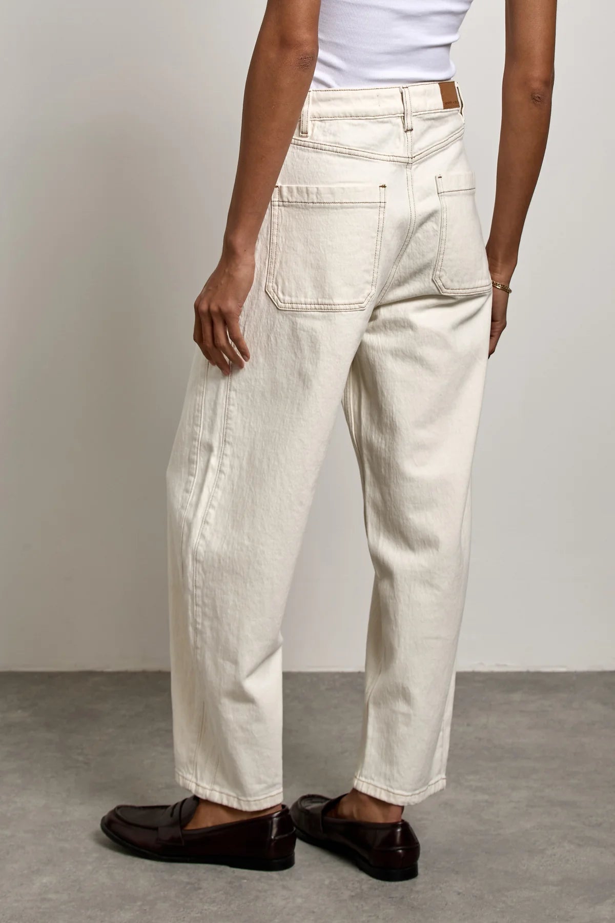 Wearing Seventy & Mochi’s Bo Barrel Leg Jeans in ecru, paired with a white sleeveless top and dark brown loafers, the person stands on a gray floor facing away from the camera.
