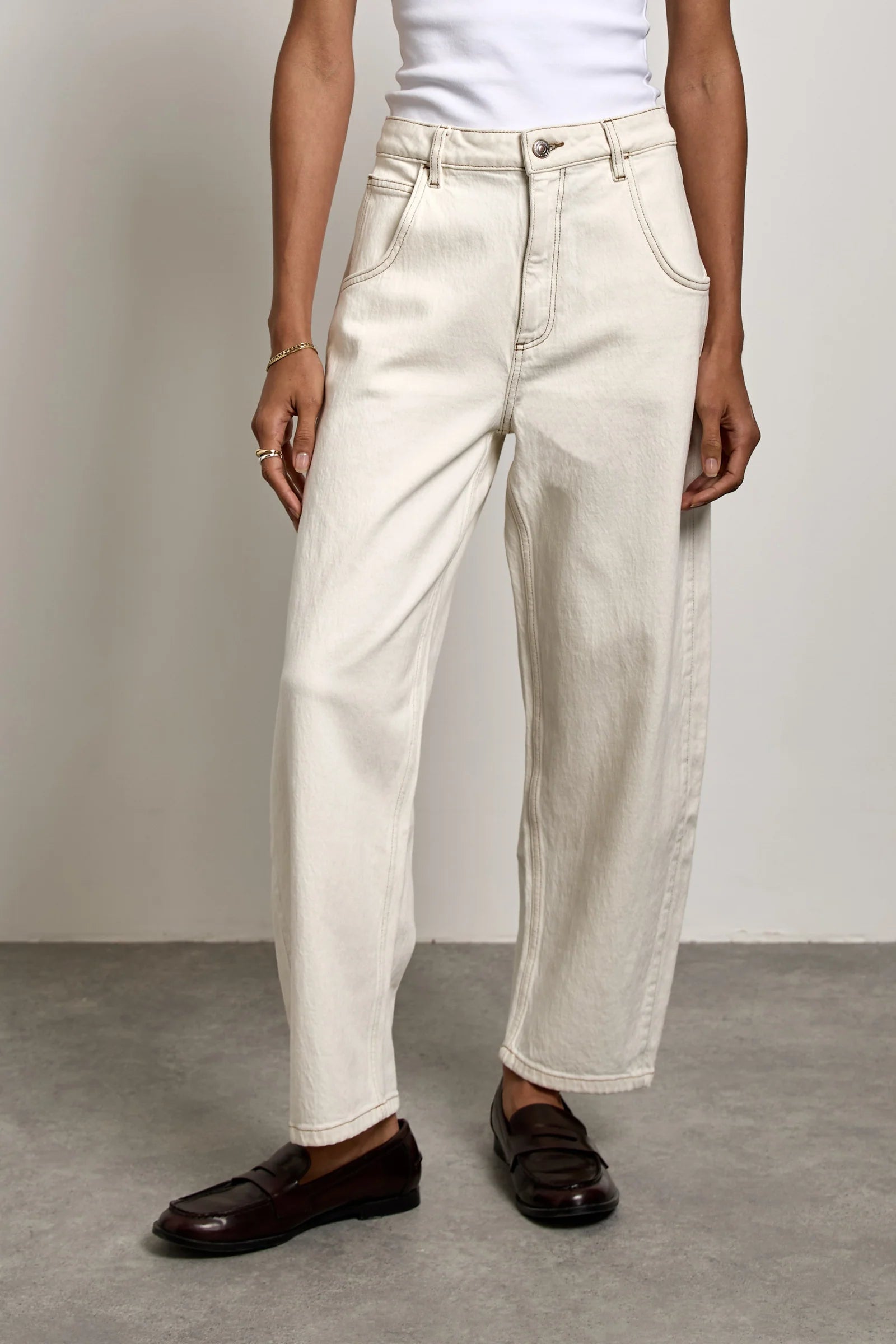 A person stands against a plain wall, showcasing the lower half of their body dressed in Seventy & Mochi’s Bo Barrel Leg Jeans in ecru—a rigid denim style featuring a super high-rise waist—paired with a white top and dark loafers.