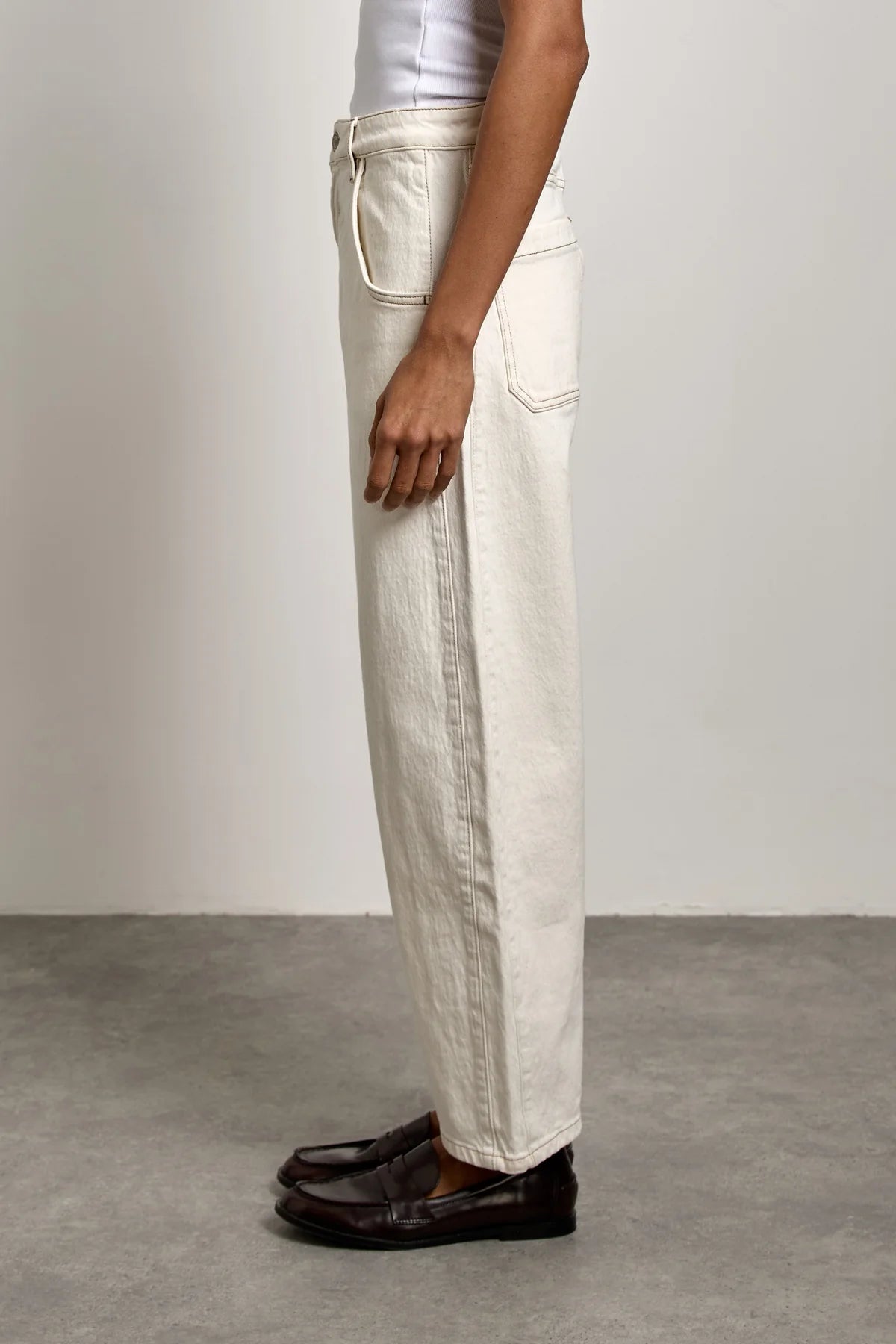 A person stands sideways against a plain gray floor and light background, wearing Seventy & Mochi’s Bo Barrel Leg Jeans in ecru, styled with a white top and dark brown loafers.