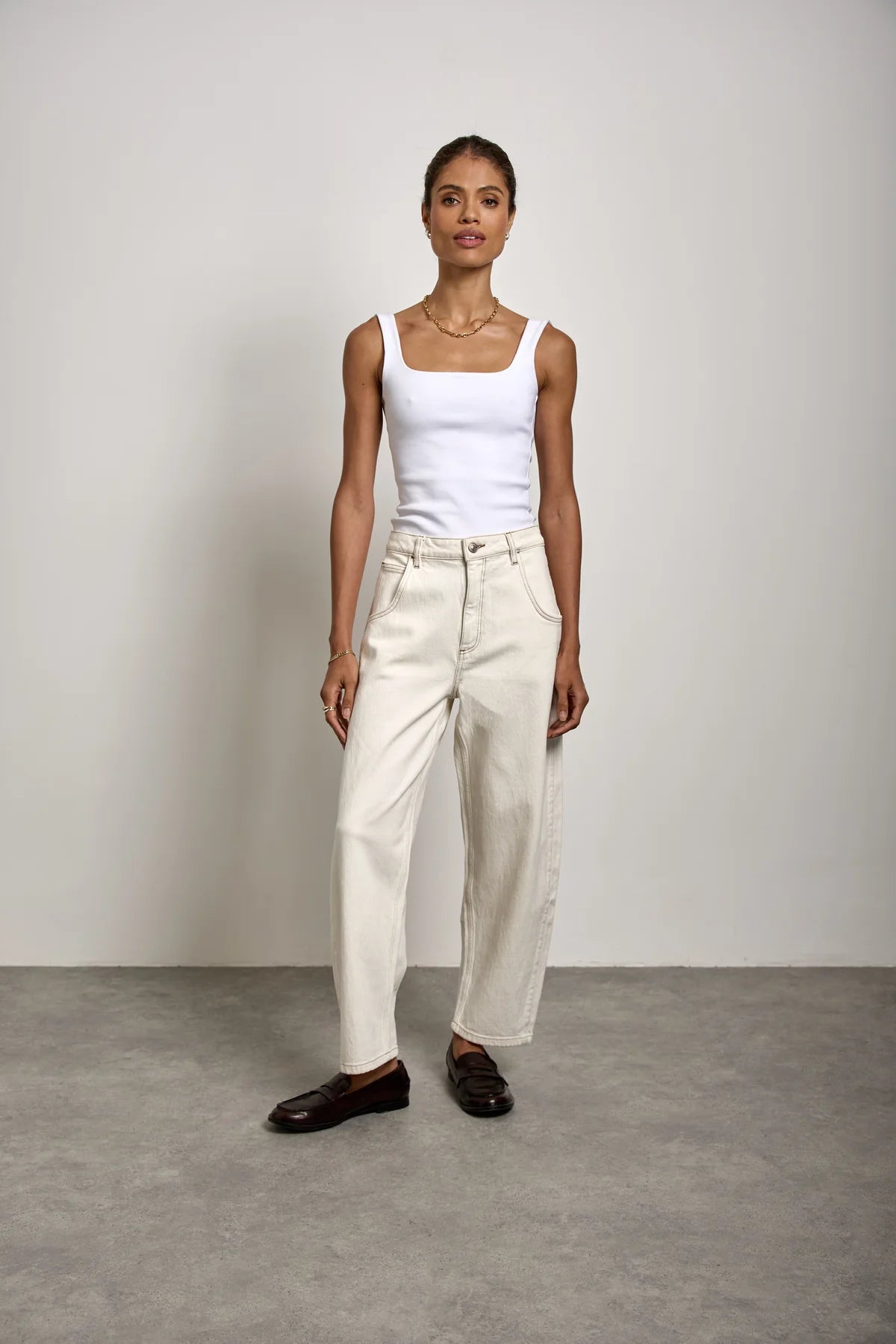 A woman stands against a plain background wearing a white tank top, Seventy & Mochi’s Bo Barrel Leg Jeans in ecru with a super high-rise waist and barrel leg silhouette, paired with dark loafers.