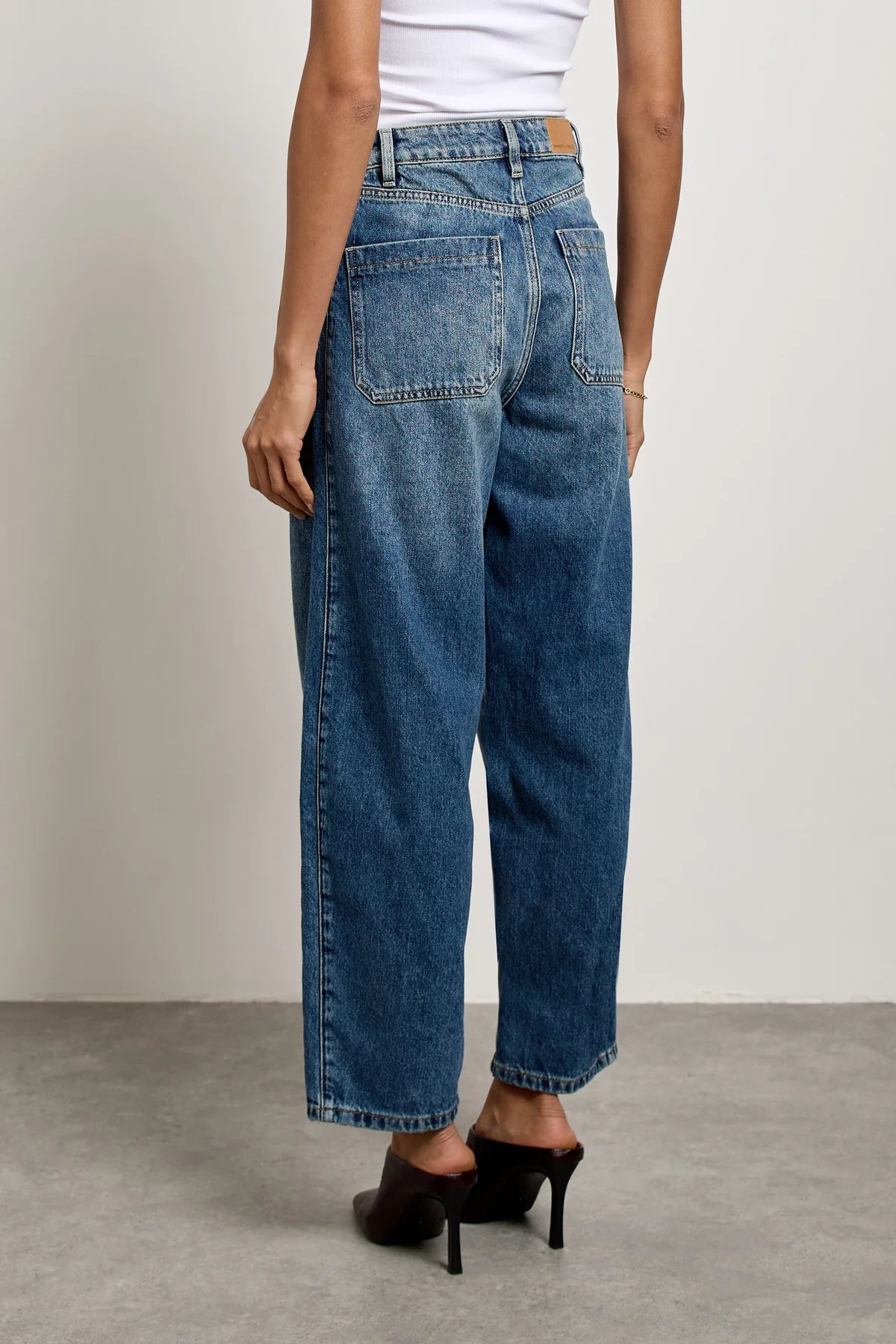 Model wearing Seventy & Mochi’s Bo Barrel Leg Jeans in Phoenix, paired with black heels, photographed from behind against a plain light wall and gray floor.
