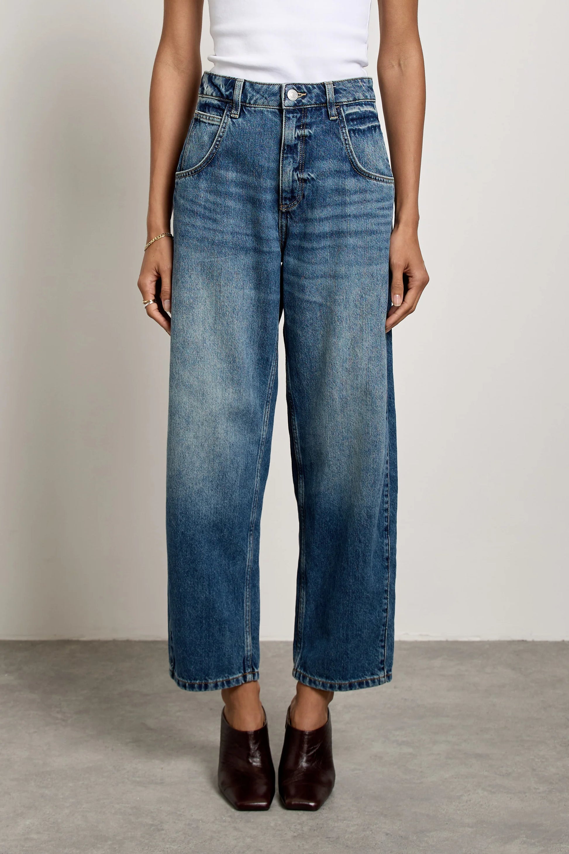 A person wearing Seventy & Mochi Bo Barrel Leg Jeans - Phoenix, featuring a super high-rise and wide-leg silhouette in rigid blue denim, paired with brown shoes, stands on a gray floor against a plain light background.