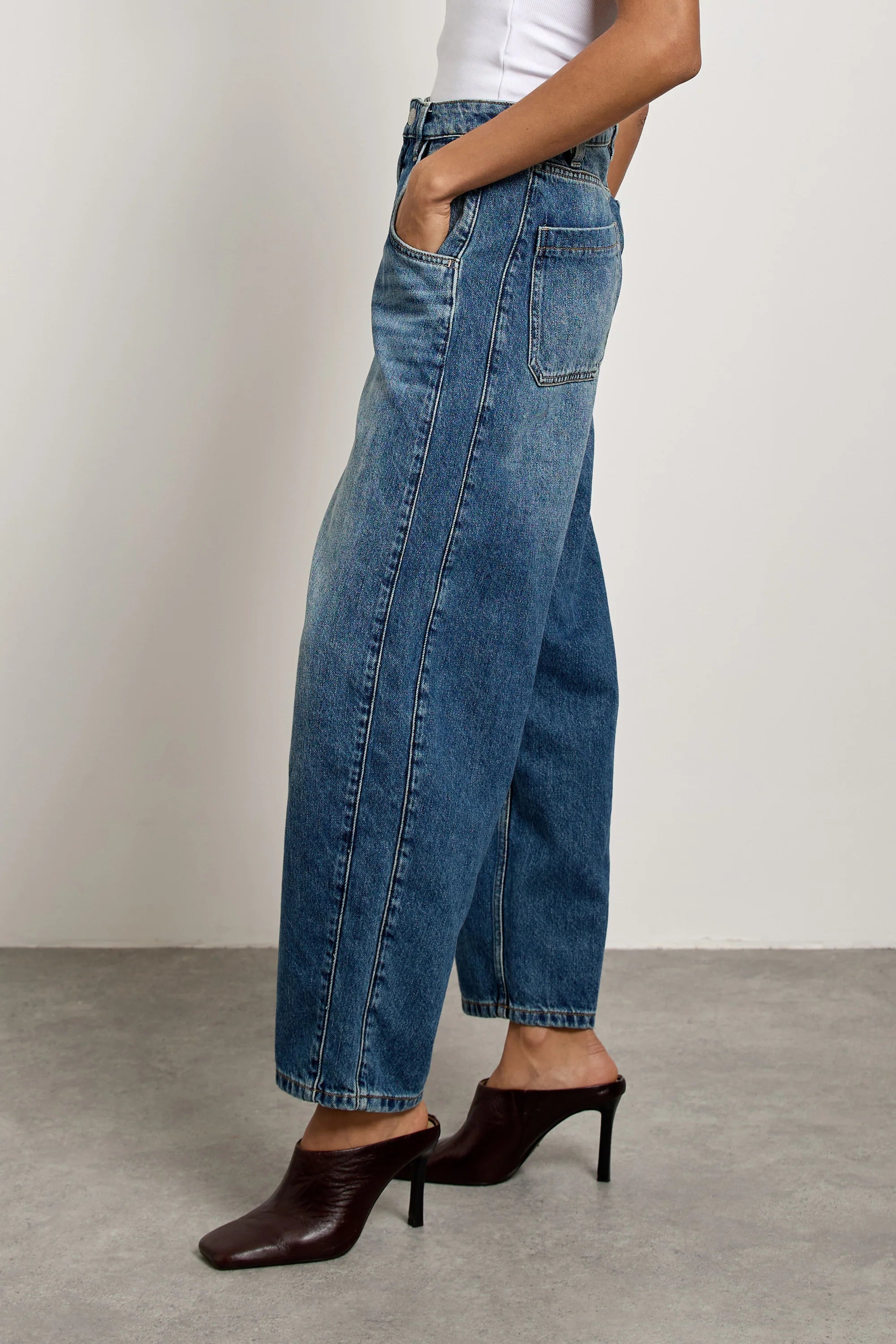 A person wearing Seventy & Mochi’s Bo Barrel Leg Jeans - Phoenix, paired with a white top and brown heeled mules, stands on a gray floor with hands in pockets against a plain light-colored wall.