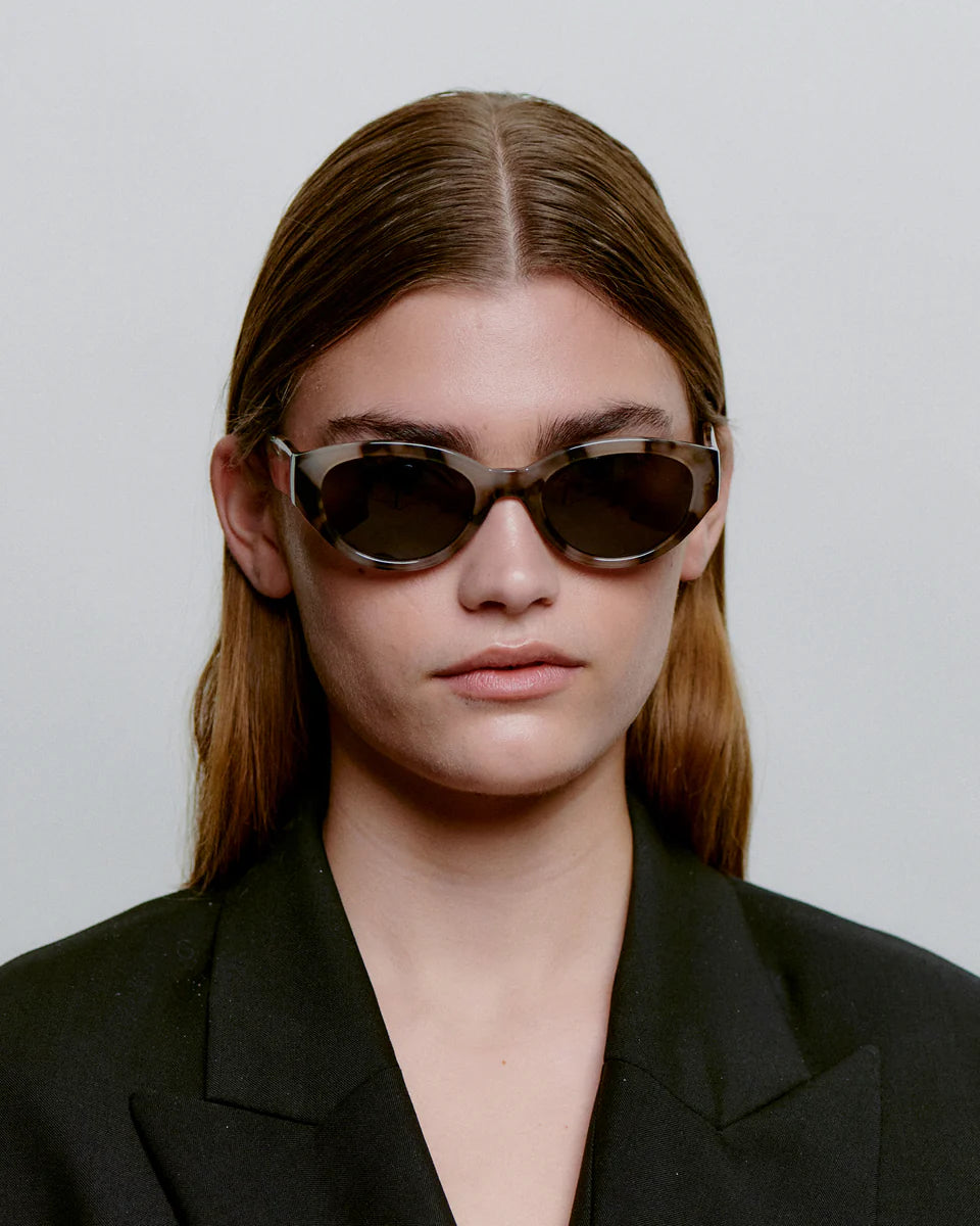 A person with straight brown hair wears A. Kjærbede's Winnie Sunglasses in Cookie Dough and a black blazer, capturing Scandinavian style against a simple light background.