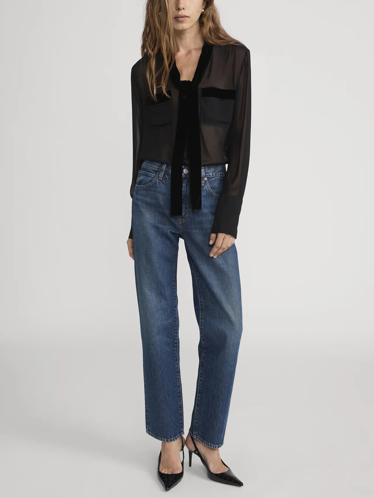 A woman stands against a plain background, wearing Frame's The Hang - Sail: a sheer black blouse with front pockets, relaxed rigid denim jeans, and black pointed-toe heels.