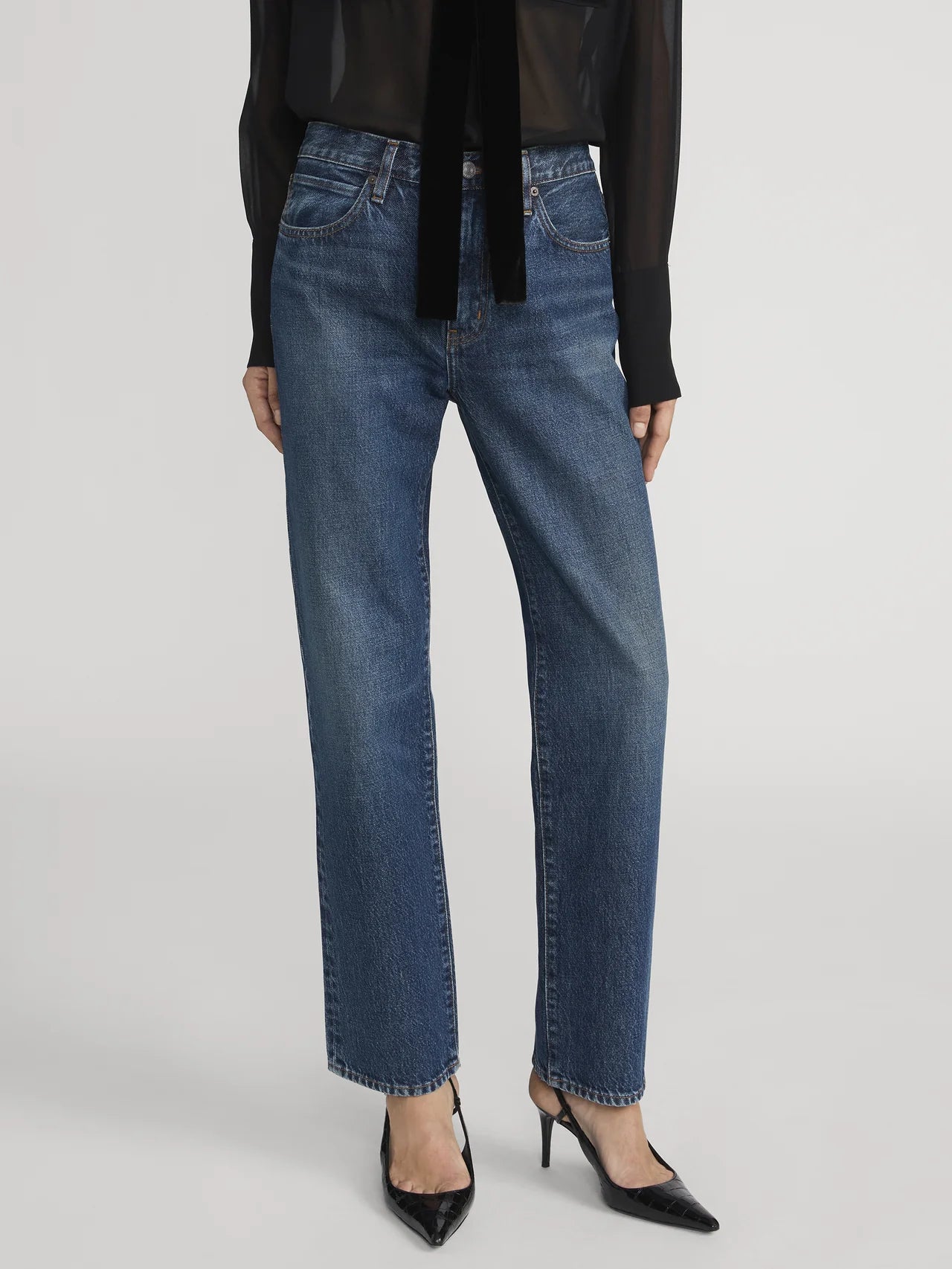 Person wearing Frame's The Hang - Sail relaxed mid-rise straight-leg blue jeans in rigid denim, a sheer black long-sleeve blouse, and black pointed-toe heels, standing against a plain background.