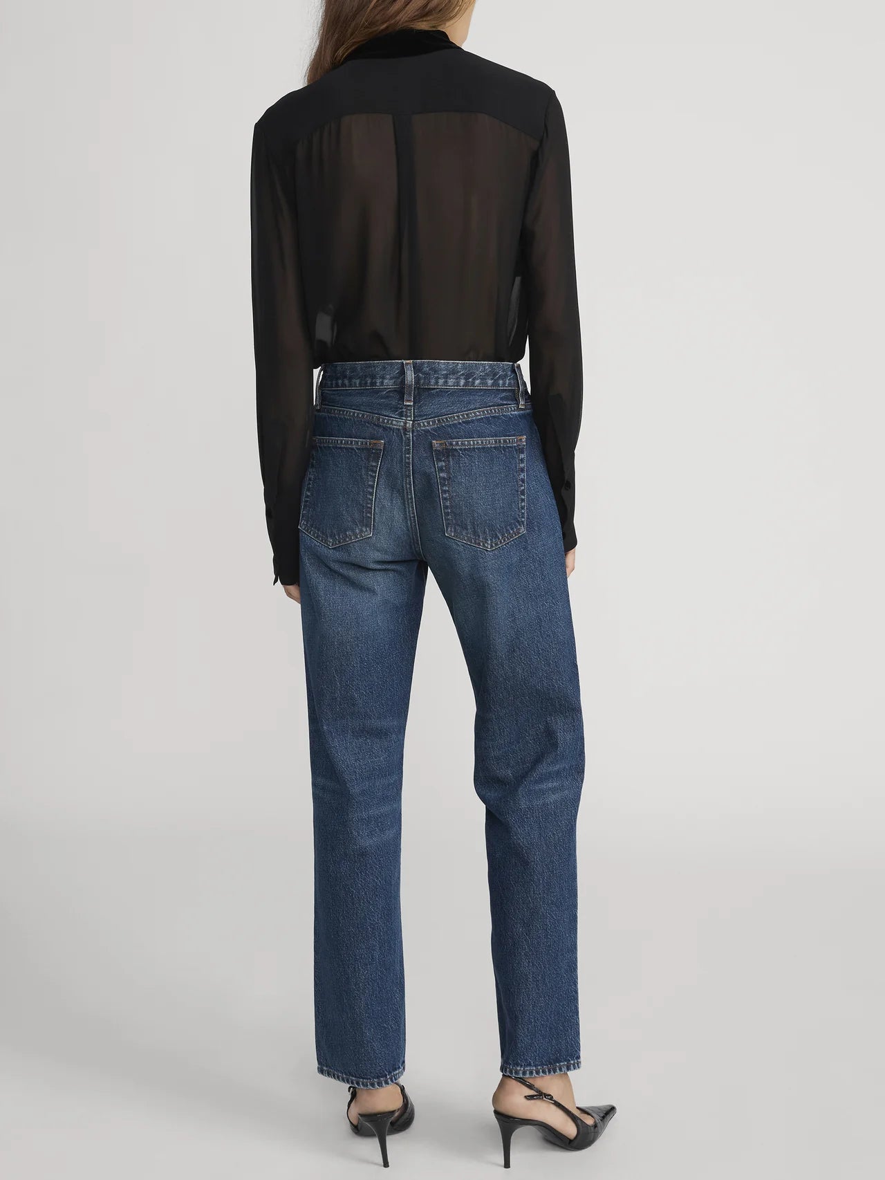 A person stands with their back to the camera, wearing Frame’s The Hang - Sail relaxed high-waisted blue jeans made from regenerative cotton, a sheer black long-sleeve shirt, and black heeled shoes.