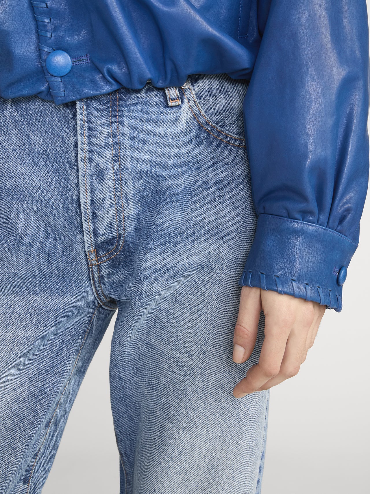 A person wearing Frame's The Essential - Rapture light blue boyfriend jeans made from regenerative cotton, paired with a blue leather jacket with stitched cuffs, shown waist to mid-thigh with one hand visible.
