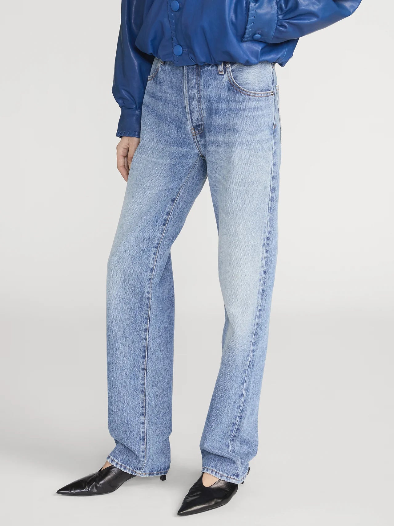 Wearing The Essential - Rapture light blue boyfriend jeans by Frame, paired with a blue long-sleeve shirt and black pointed-toe shoes, this person stands against a plain background.