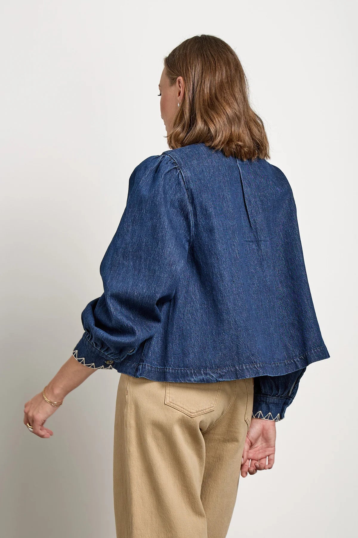 A woman with shoulder-length brown hair, wearing the Seventy & Mochi Ziggy Jacket - Americana in a relaxed fit dark blue organic cotton denim and beige pants, stands with her back to the camera against a plain background.