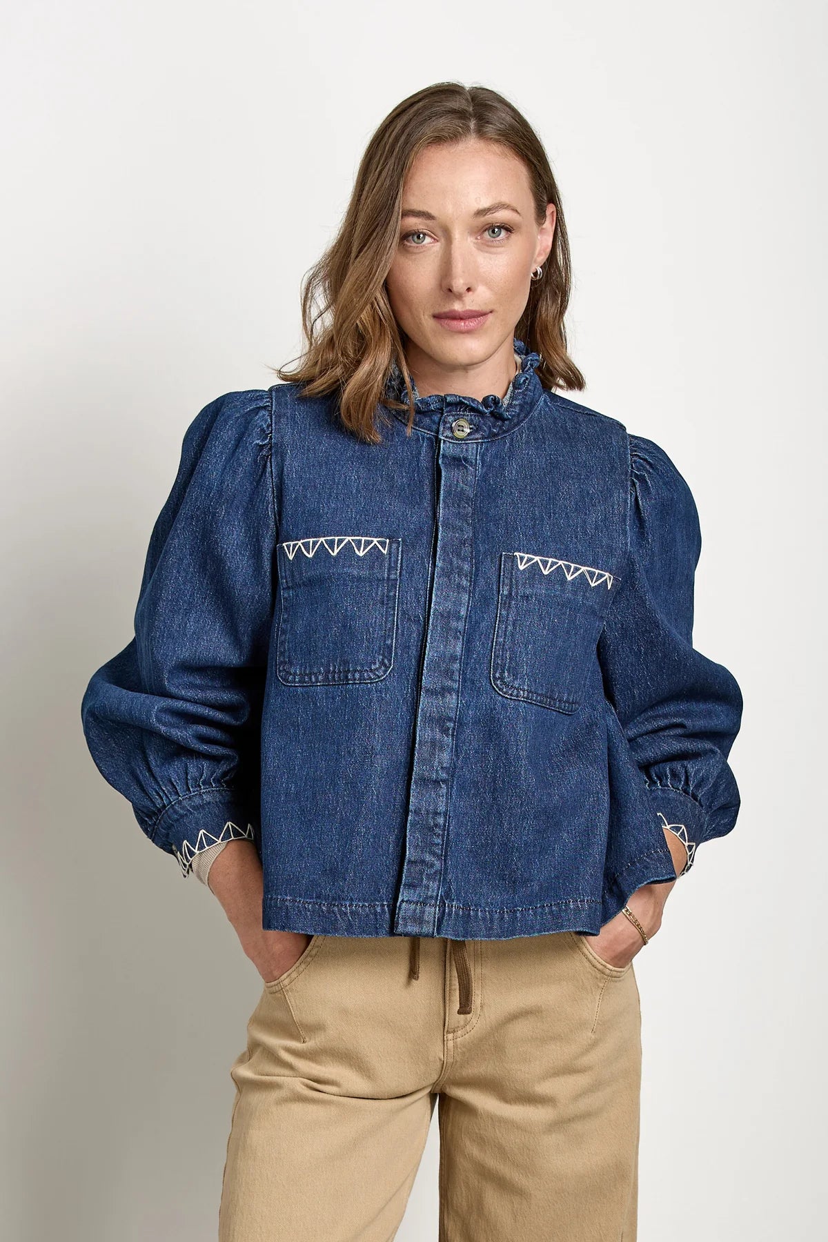 A woman stands with her hands in her pockets against a plain background, wearing Seventy & Mochi’s Ziggy Jacket - Americana, a relaxed fit denim piece with puffed sleeves, paired with tan organic cotton pants.