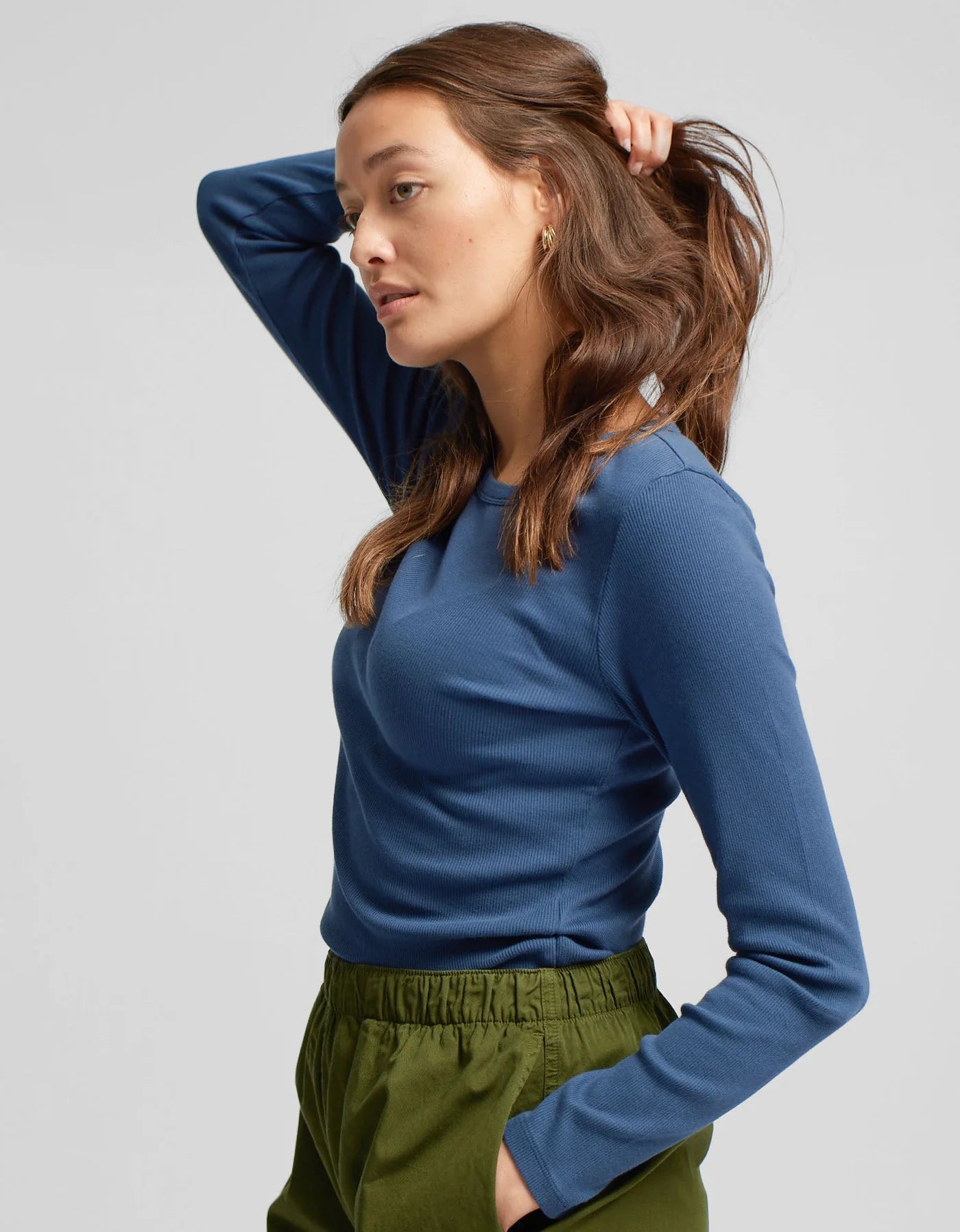 A woman is standing, wearing a blue Organic Rib LS T-Shirt by Colorful Standard made of organic cotton and green pants. She has her right hand in her hair and is looking to the side.
