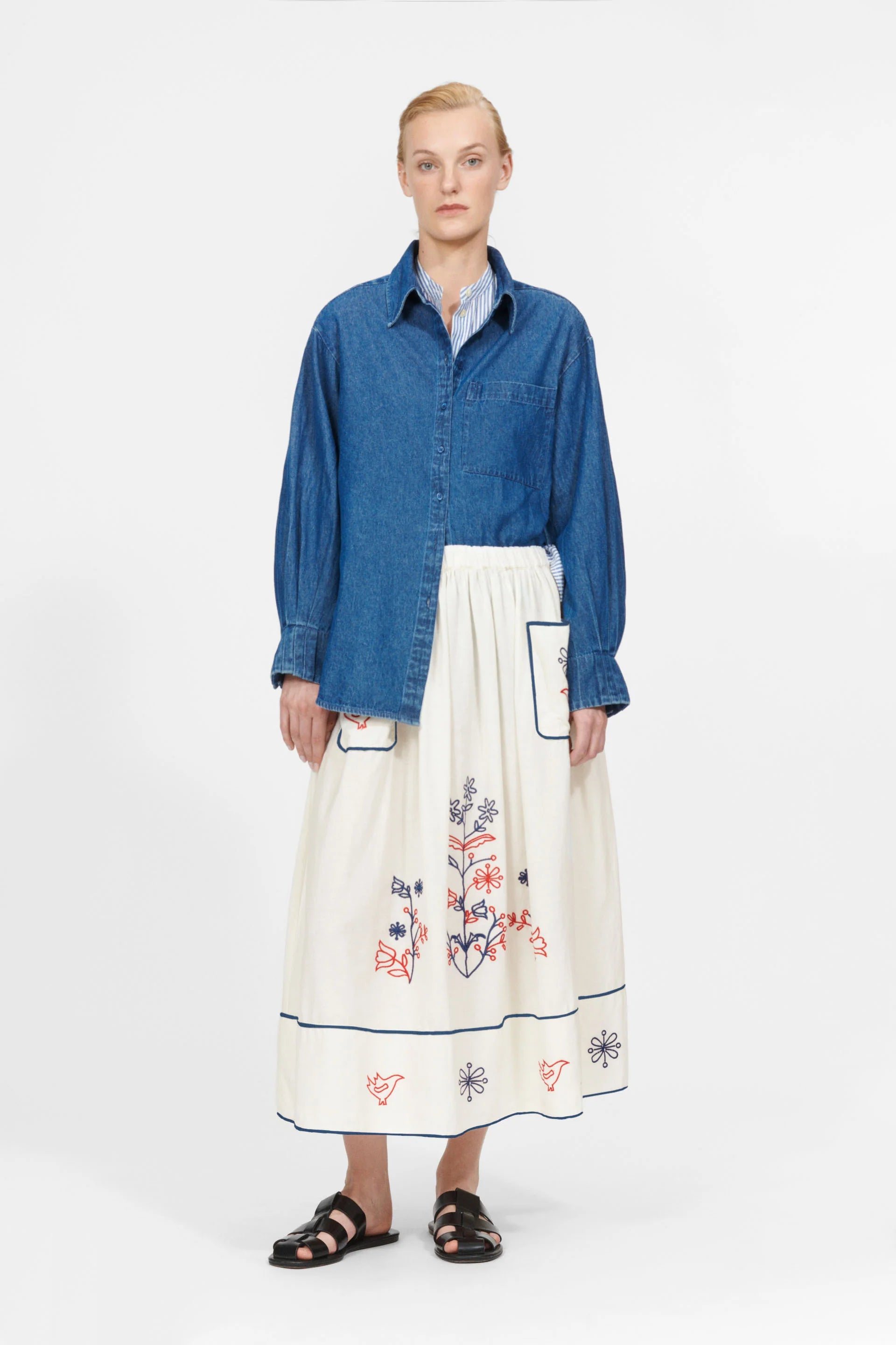 A person stands on a plain background wearing the Nué Notes York Shirt in blue denim, paired with a white skirt featuring colorful embroidery for a modern twist.