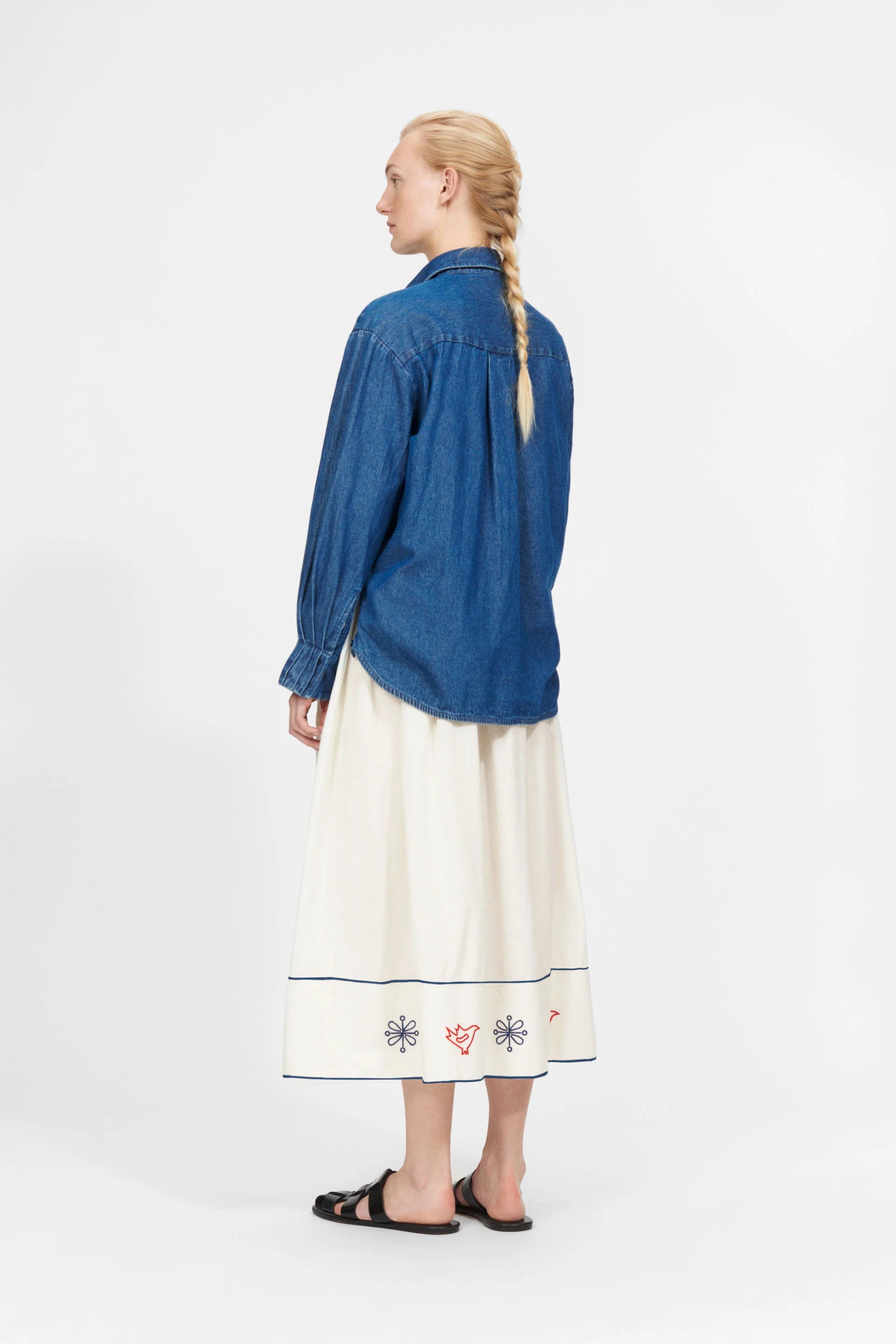 A person with a long braid stands against a plain backdrop, wearing the oversized Nué Notes York Shirt in blue denim, paired with a white skirt adorned with floral embroidery and black sandals, adding a modern twist to their style.