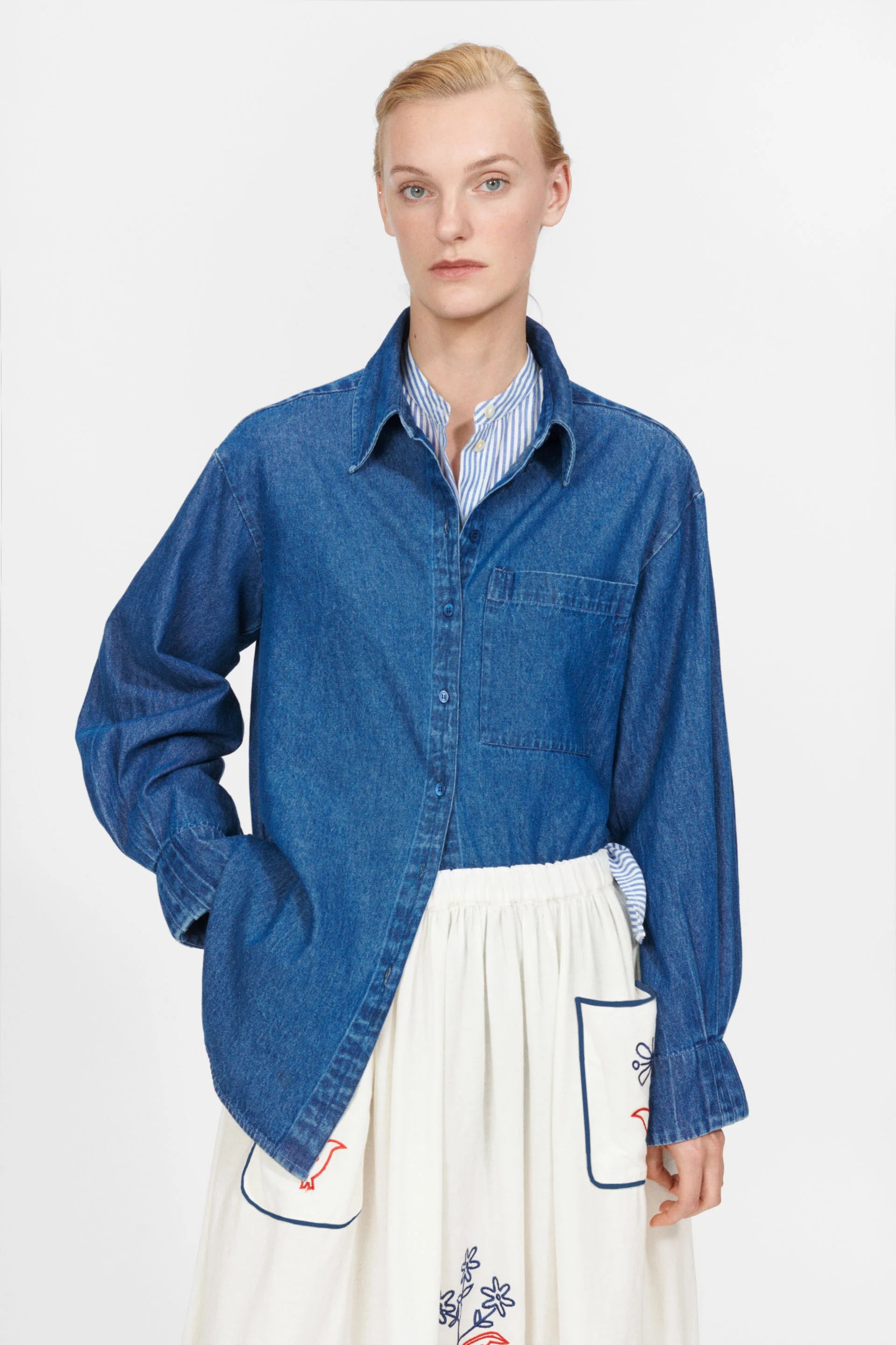 Wearing the York Shirt in blue denim by Nué Notes over a striped blouse and a white skirt with decorative pockets, the person stands elegantly against a plain background, adding a modern twist to the ensemble.