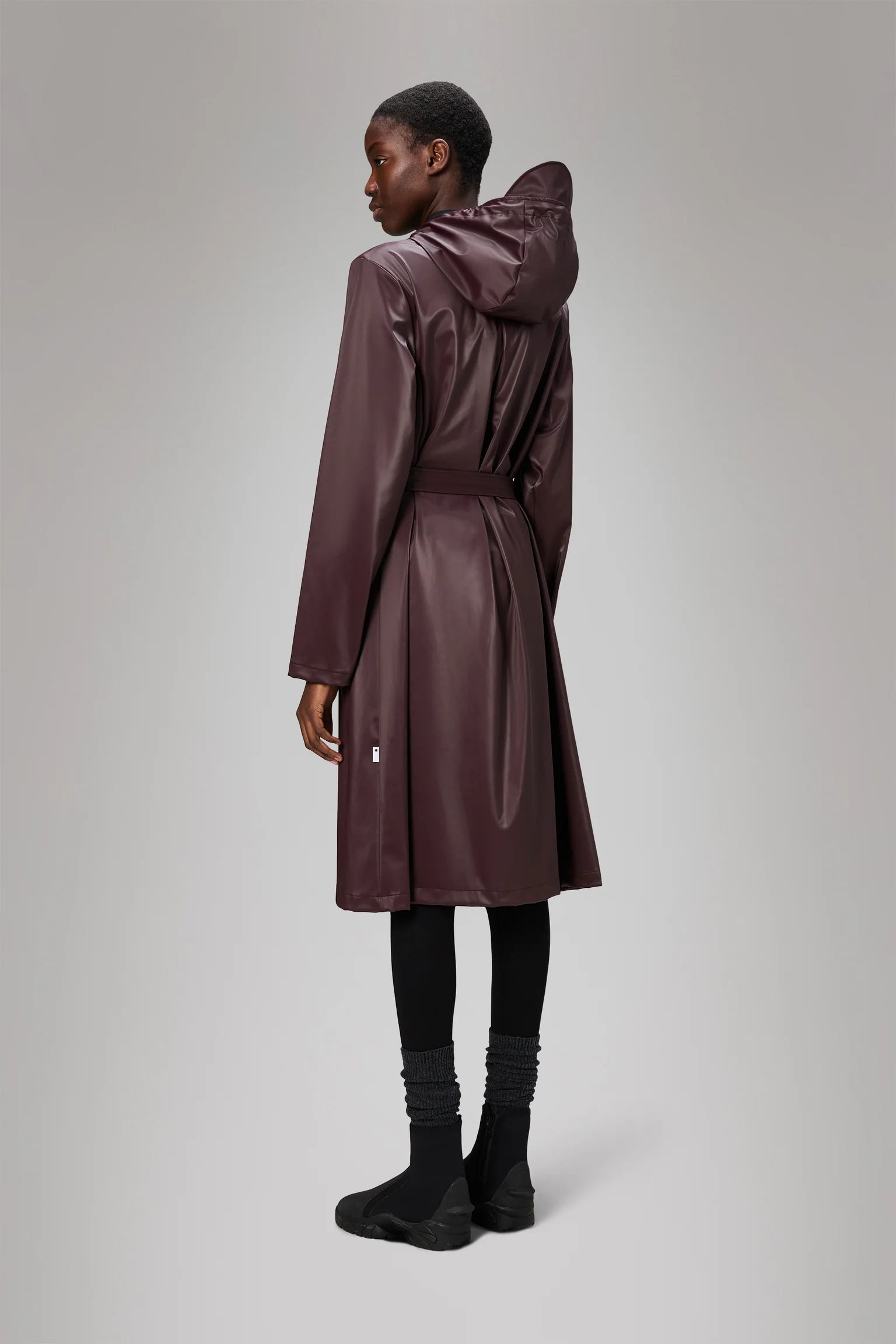 A person stands facing away, wearing the Rains A-Line Longer W Jacket - Depth, a long dark brown hooded raincoat, with black leggings and shoes against a plain light gray background.