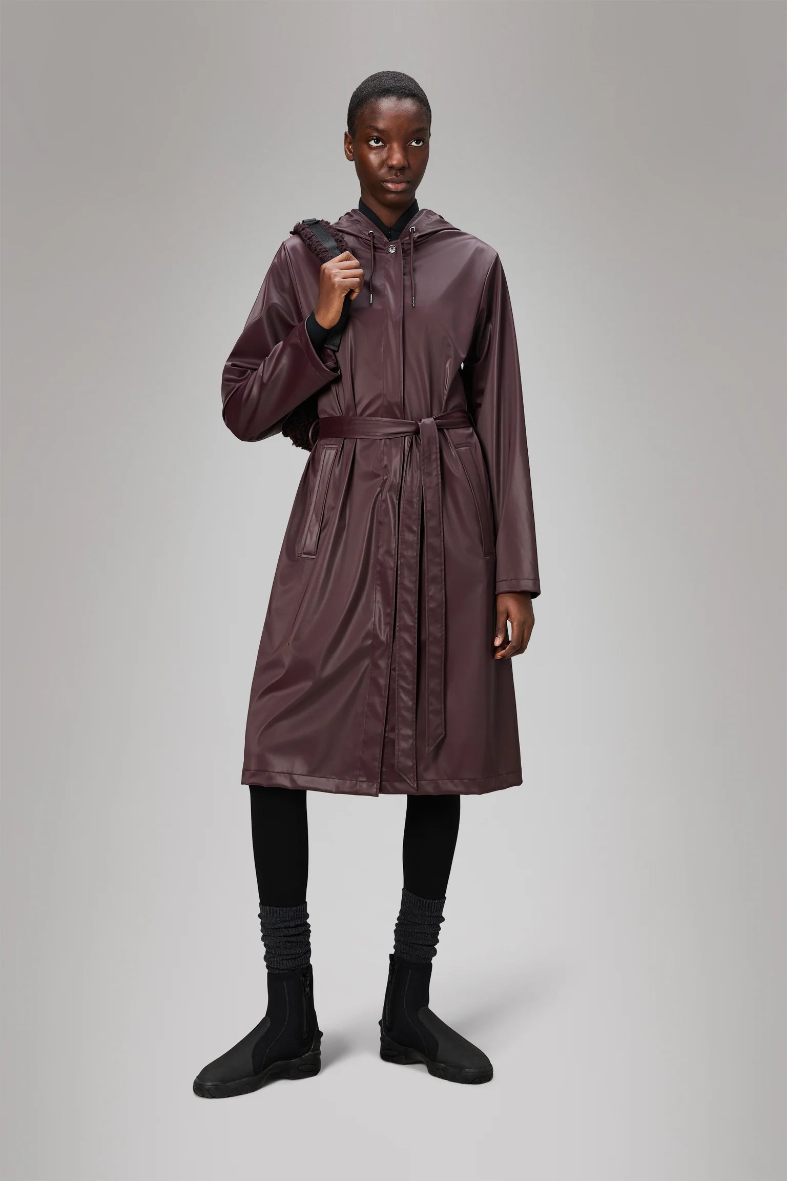 A person stands against a plain background wearing the Rains A-Line Longer W Jacket – Depth, a long belted maroon raincoat with hood, paired with black leggings, socks, ankle boots, and carrying a bag over one shoulder.