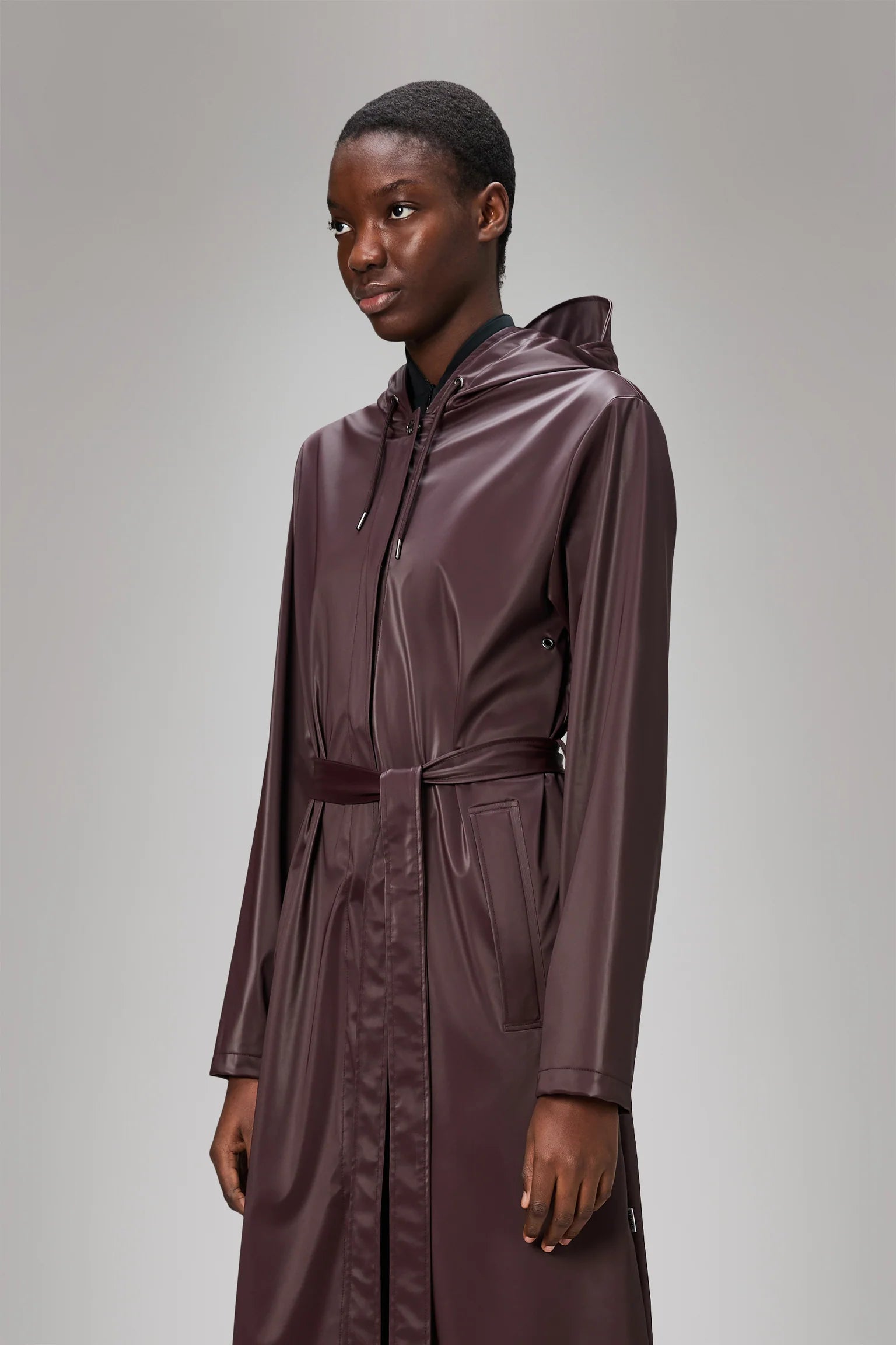 Model wears the A-Line Longer W Jacket - Depth by Rains, a burgundy women's raincoat with hood and belt, styled in an A-line silhouette, standing against a plain light gray background.