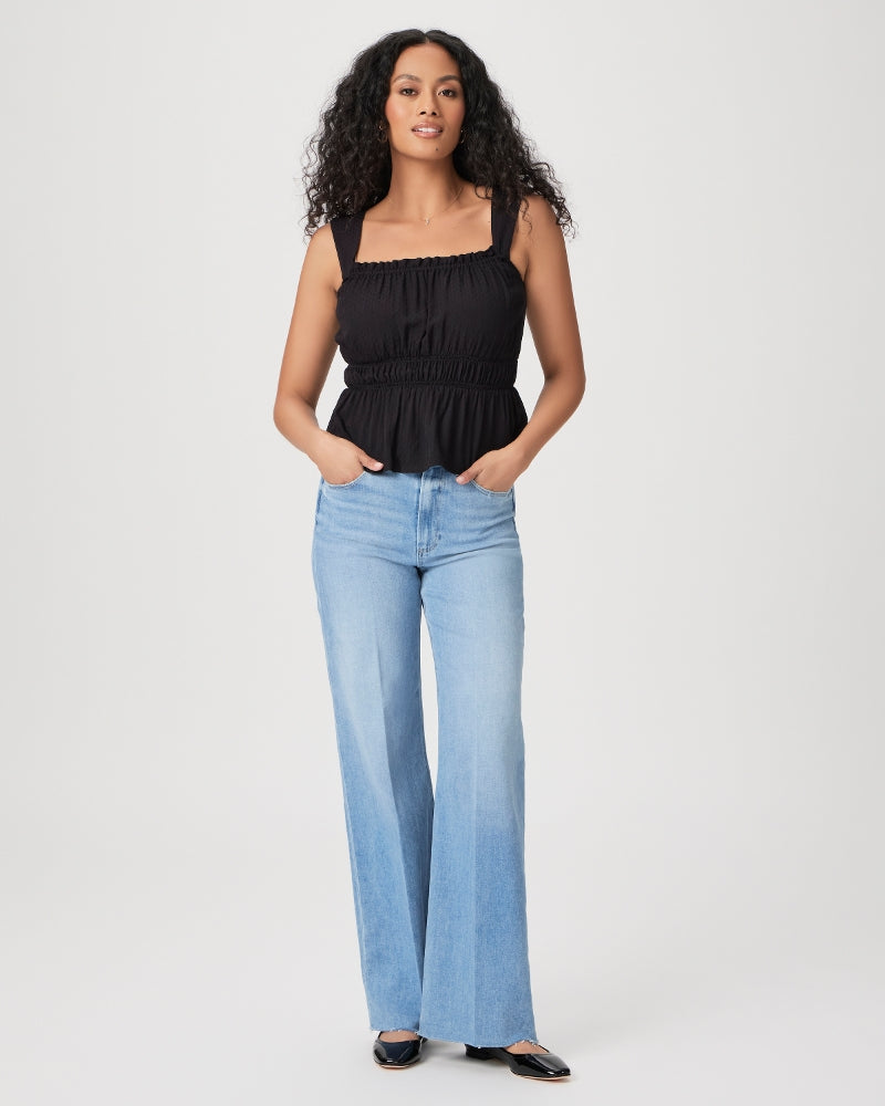 A woman standing posing in a black tank top and high-waisted blue Paige denim jeans.